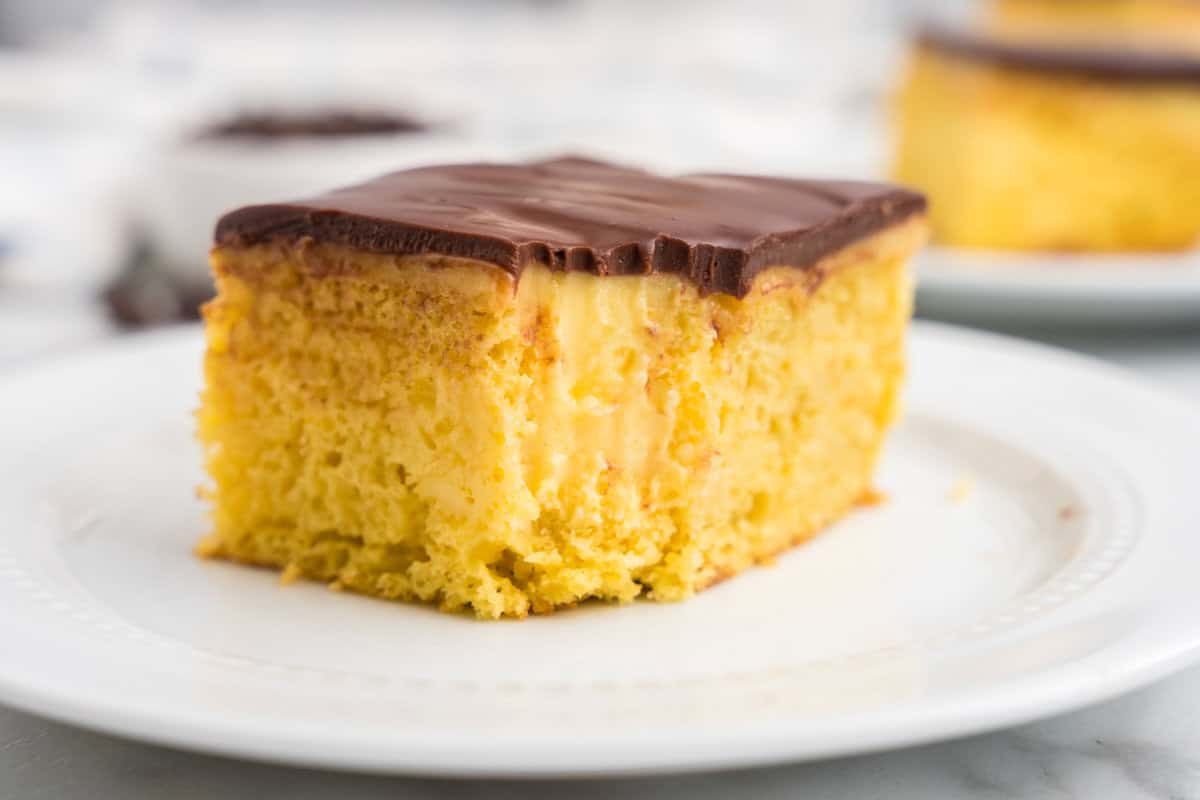 A close-up of a single slice of Boston Cream Poke Cake on a white plate, showing distinct layers of moist yellow cake, creamy vanilla pudding filling, and a thick chocolate ganache topping with a bite taken out.