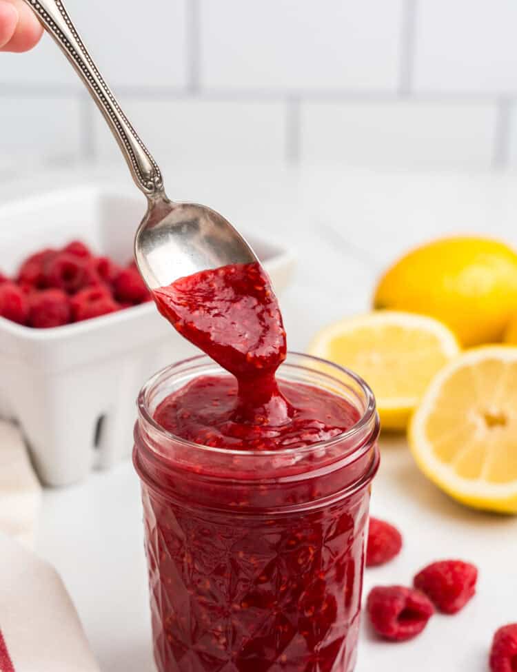 A spoon lifting thick, glossy raspberry sauce from a filled mason jar, showcasing its rich texture and deep red hue, with fresh raspberries and lemons in the background.