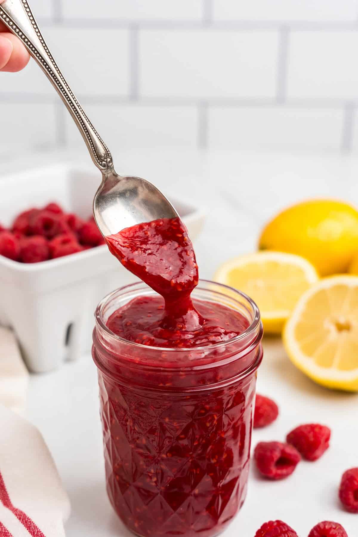 A spoon lifting thick, glossy raspberry sauce from a filled mason jar, showcasing its rich texture and deep red hue, with fresh raspberries and lemons in the background.