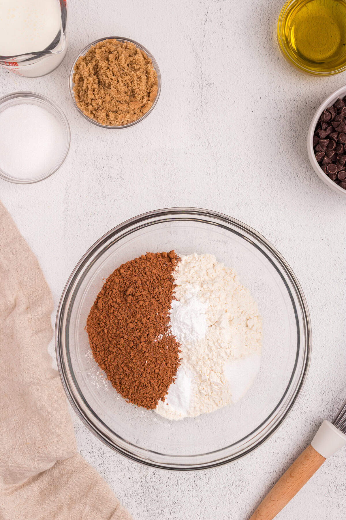A glass mixing bowl containing separated piles of cocoa powder, flour, and baking powder, surrounded by small bowls of brown sugar, white sugar, chocolate chips, and a measuring cup of milk.