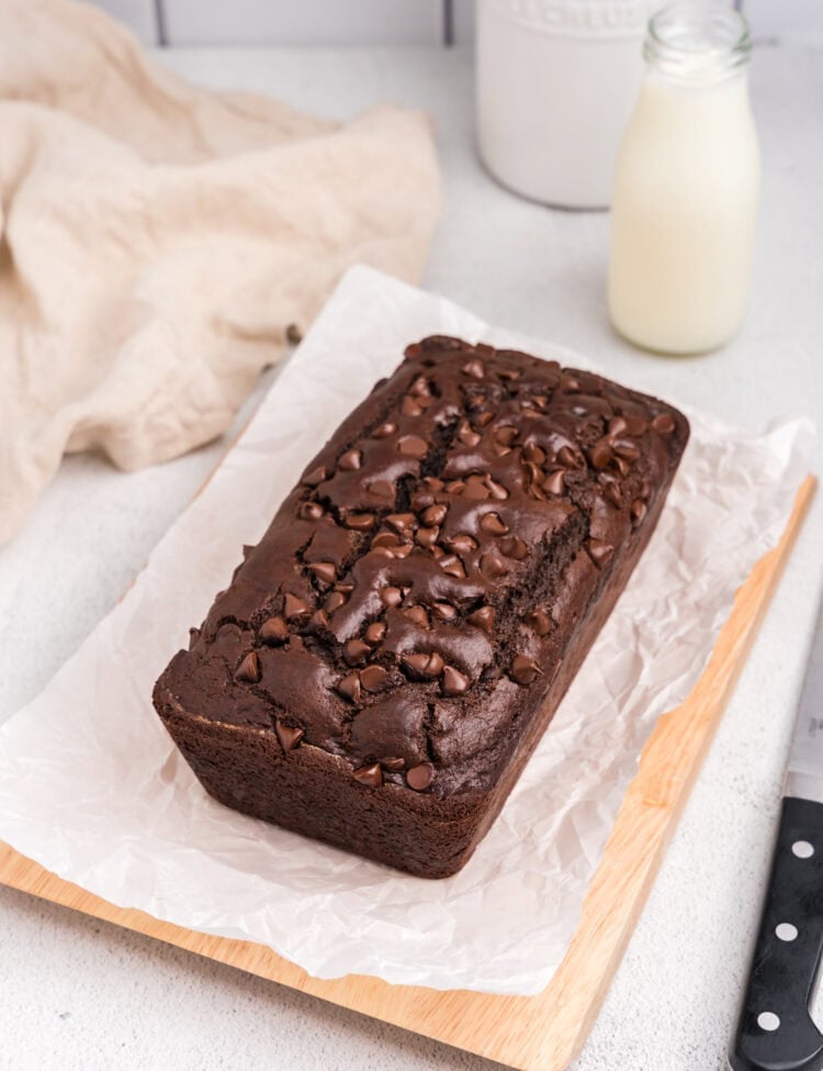 The fully baked chocolate bread placed on a parchment-lined wooden board, with a bottle of milk in the background and a soft kitchen towel beside it.