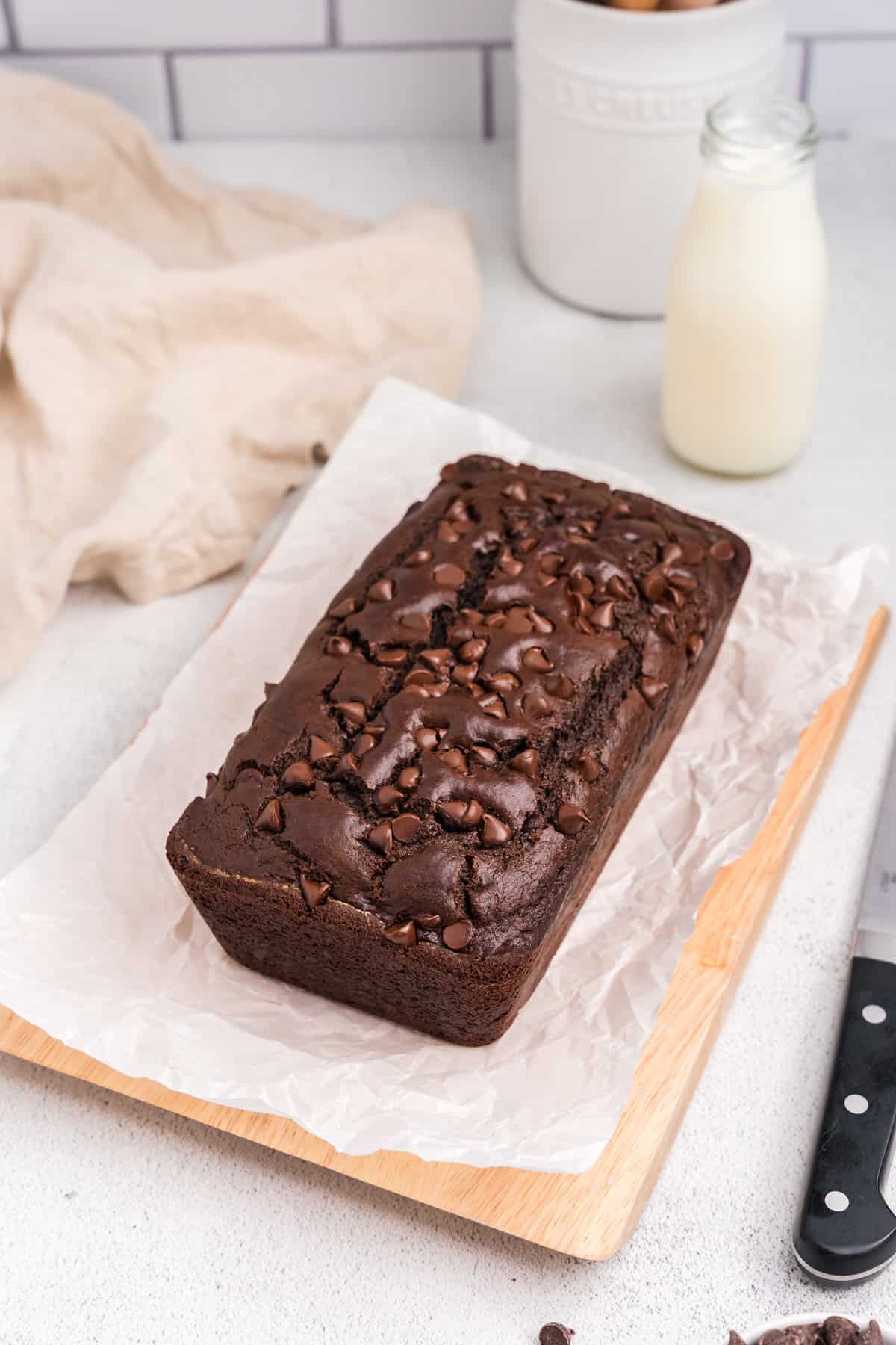 The fully baked chocolate bread placed on a parchment-lined wooden board, with a bottle of milk in the background and a soft kitchen towel beside it.