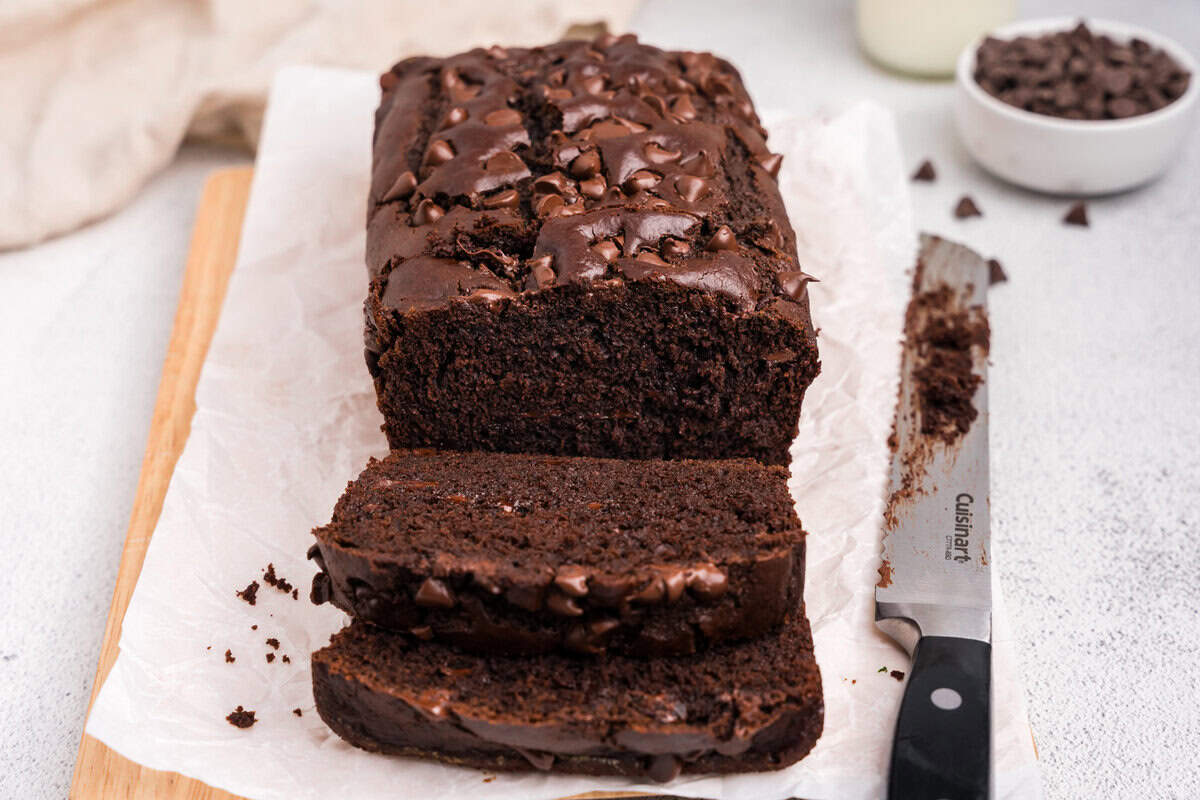 A sliced chocolate loaf on parchment paper, with two slices cut and lying in front of the loaf. A knife with chocolate crumbs on the blade rests nearby.