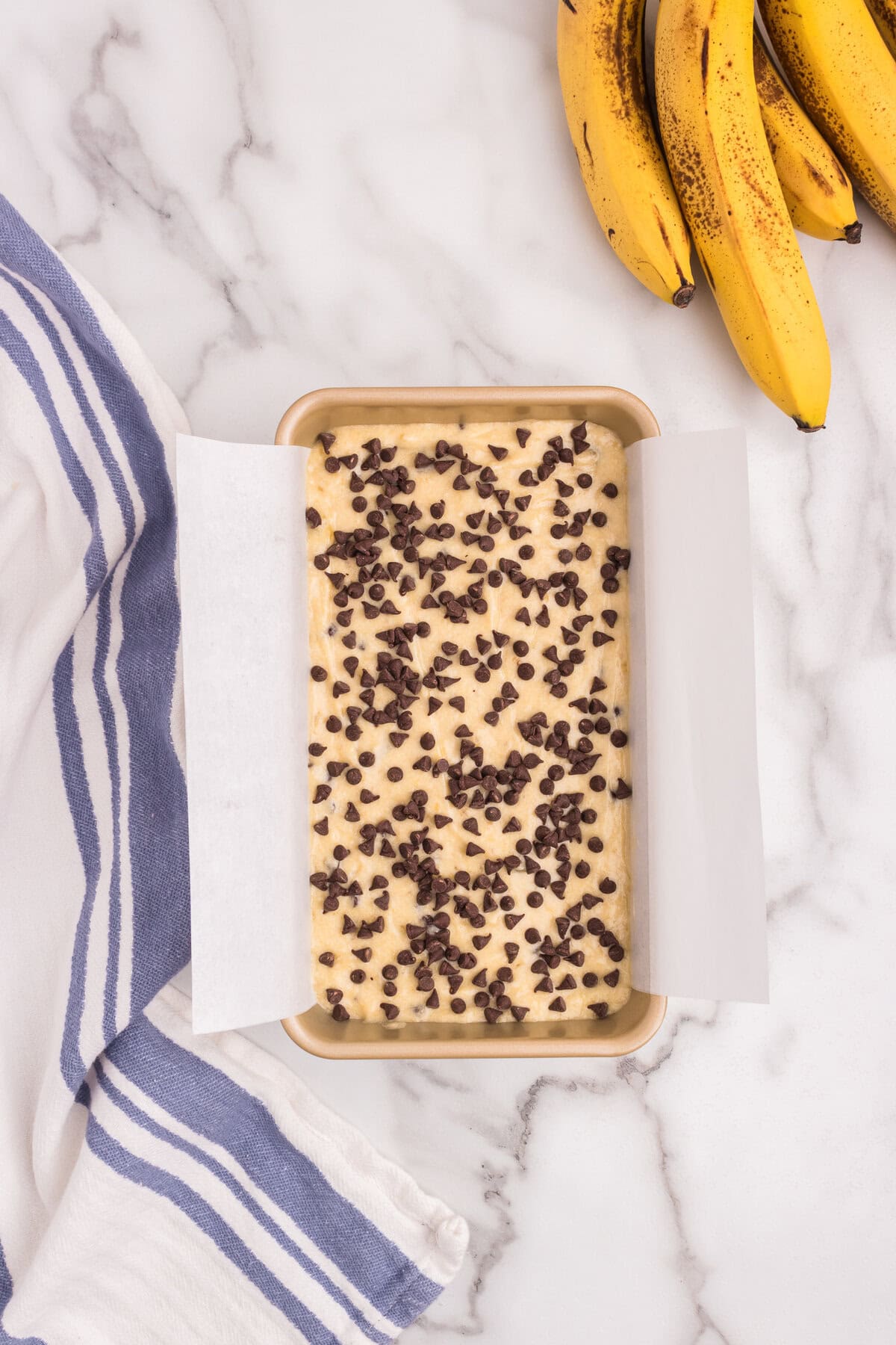 Mini chocolate chips have been sprinkled generously over the top of the batter in the loaf pan, which sits on a marble surface next to a bunch of ripe bananas.