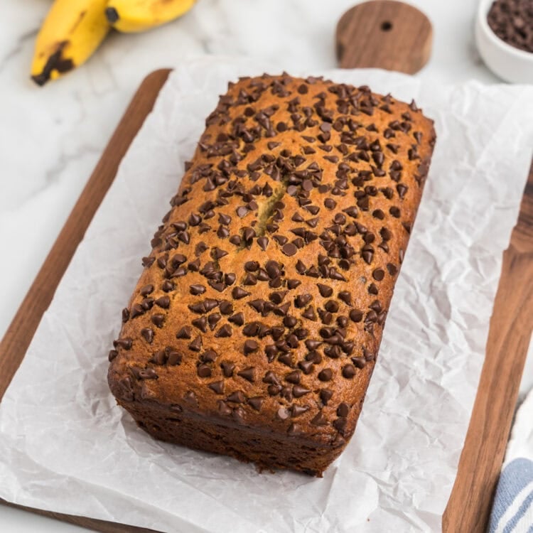 The banana bread has been removed from the pan and placed on a sheet of white parchment paper on a wooden cutting board, showing off its domed, chocolate-studded top.