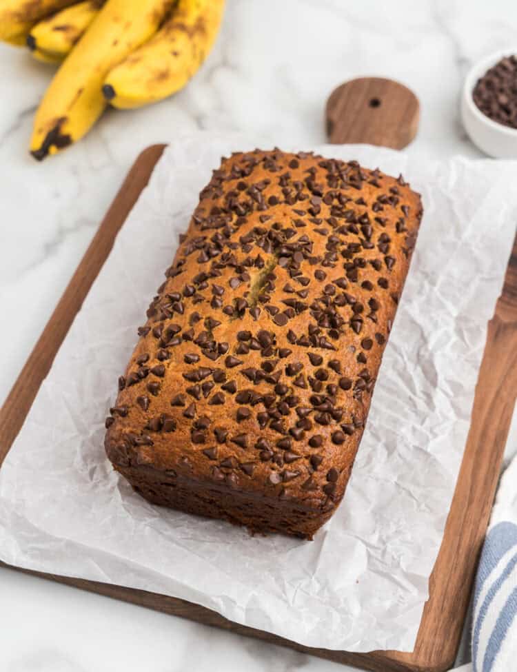 The banana bread has been removed from the pan and placed on a sheet of white parchment paper on a wooden cutting board, showing off its domed, chocolate-studded top.