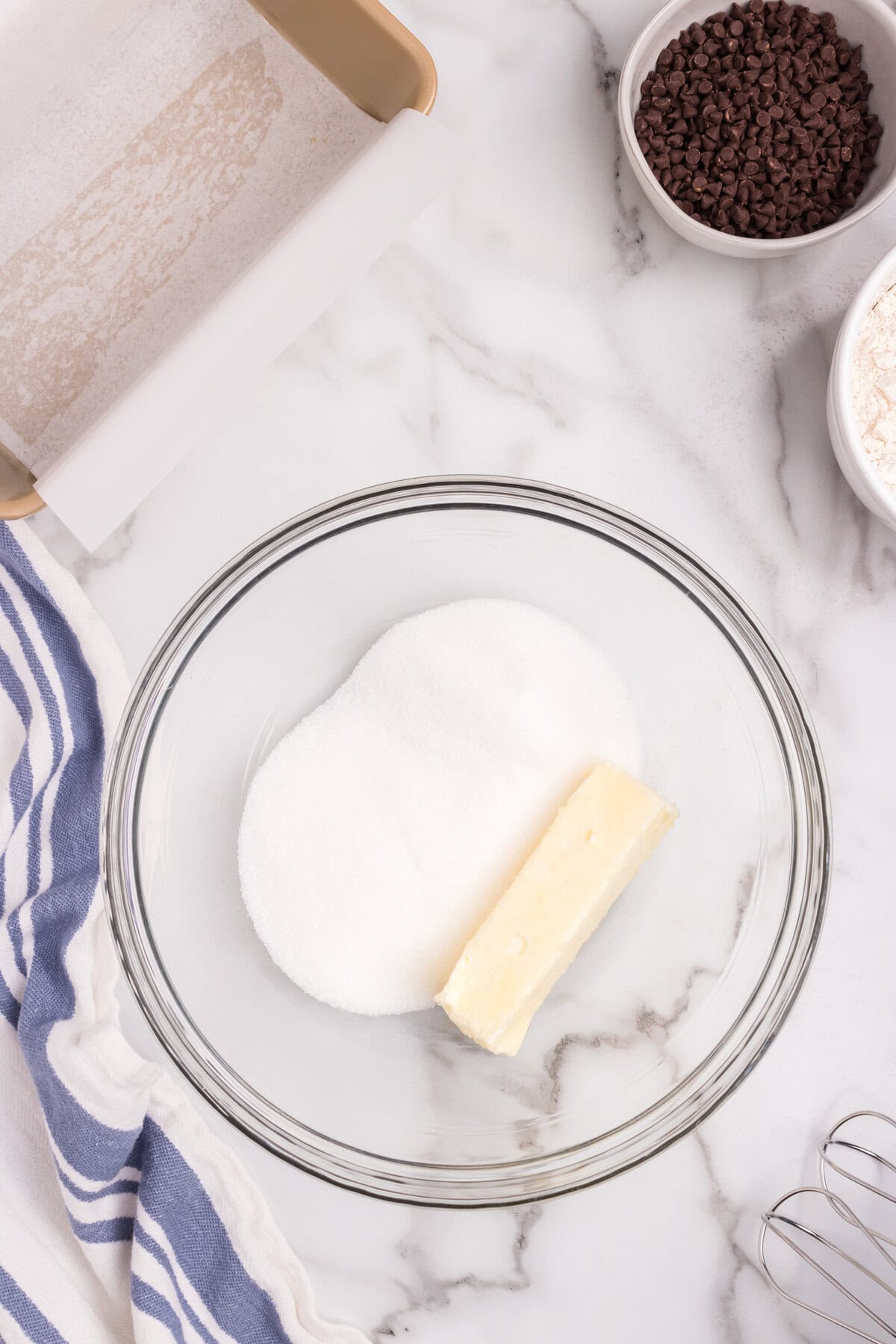 A glass mixing bowl containing a stick of butter and a mound of white sugar, ready to be creamed. The background features a loaf pan lined with parchment paper, and bowls of mini chocolate chips and flour.