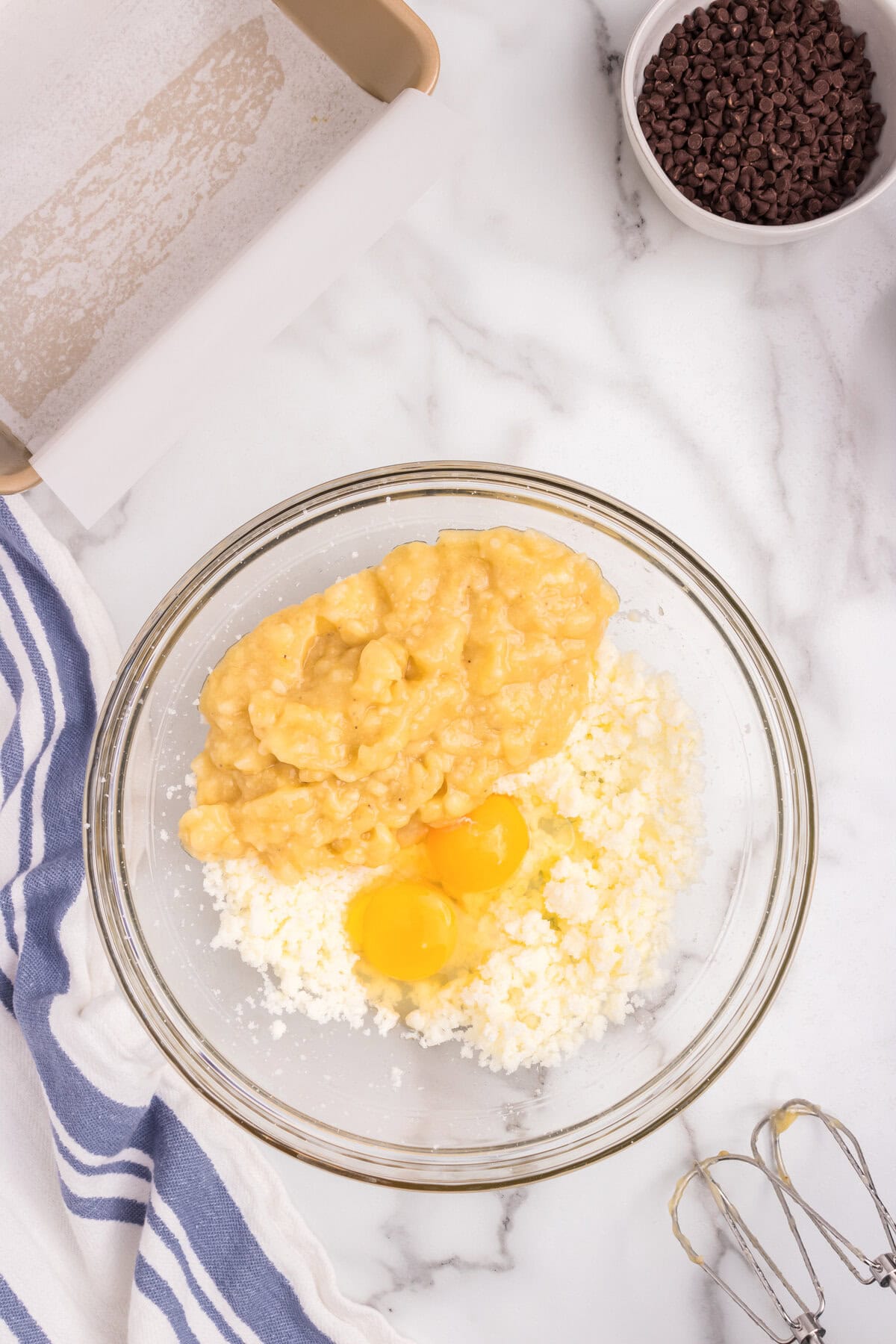 Two eggs and mashed bananas have been added to the creamed butter and sugar mixture in the bowl, ready to be mixed. The bowl rests on a marble surface with a hand mixer and ingredients nearby.