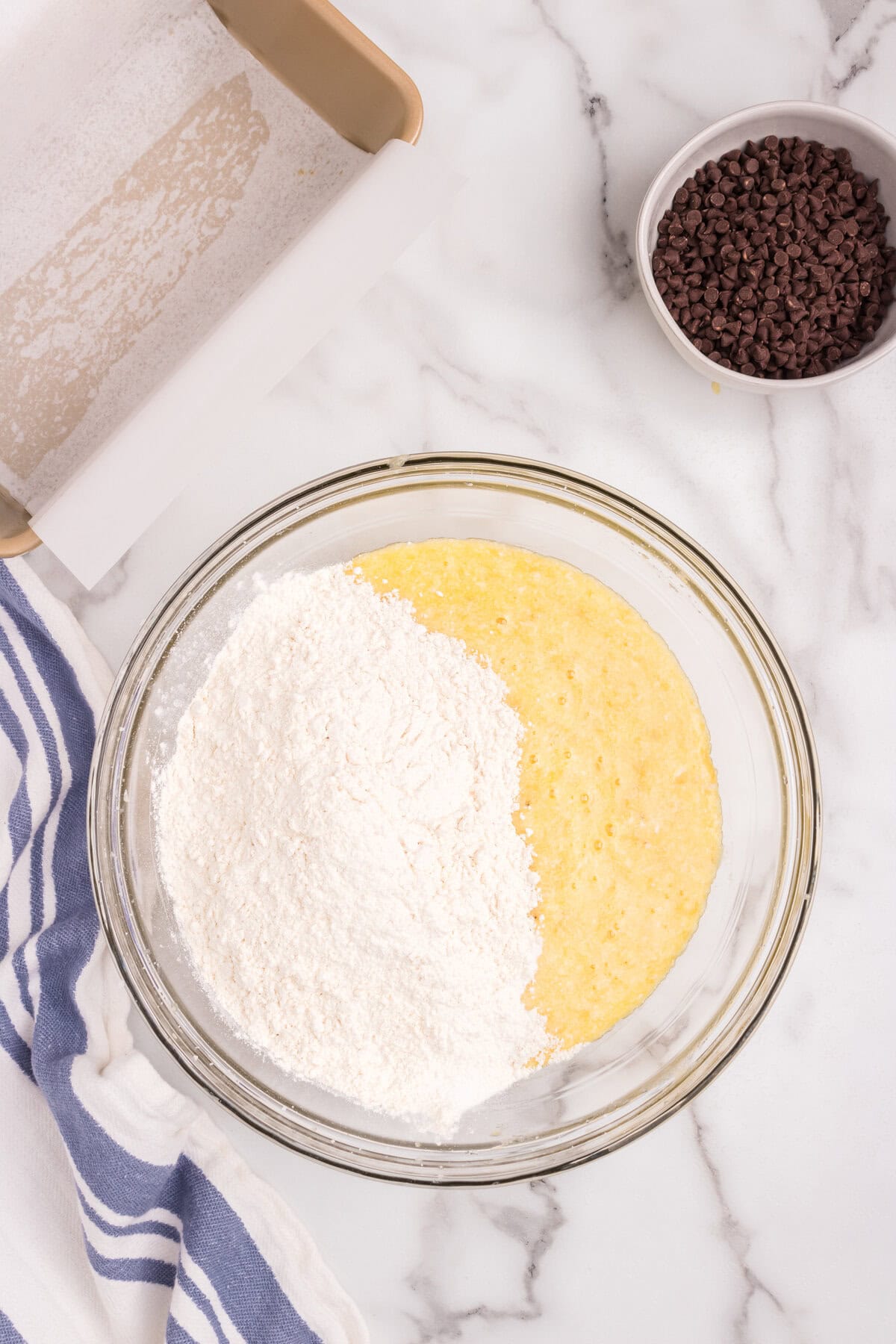 A glass mixing bowl containing partially mixed ingredients: mashed bananas and eggs on one side, and flour on the other. A gold loaf pan lined with parchment paper and a small bowl of mini chocolate chips are visible nearby on a marble countertop.