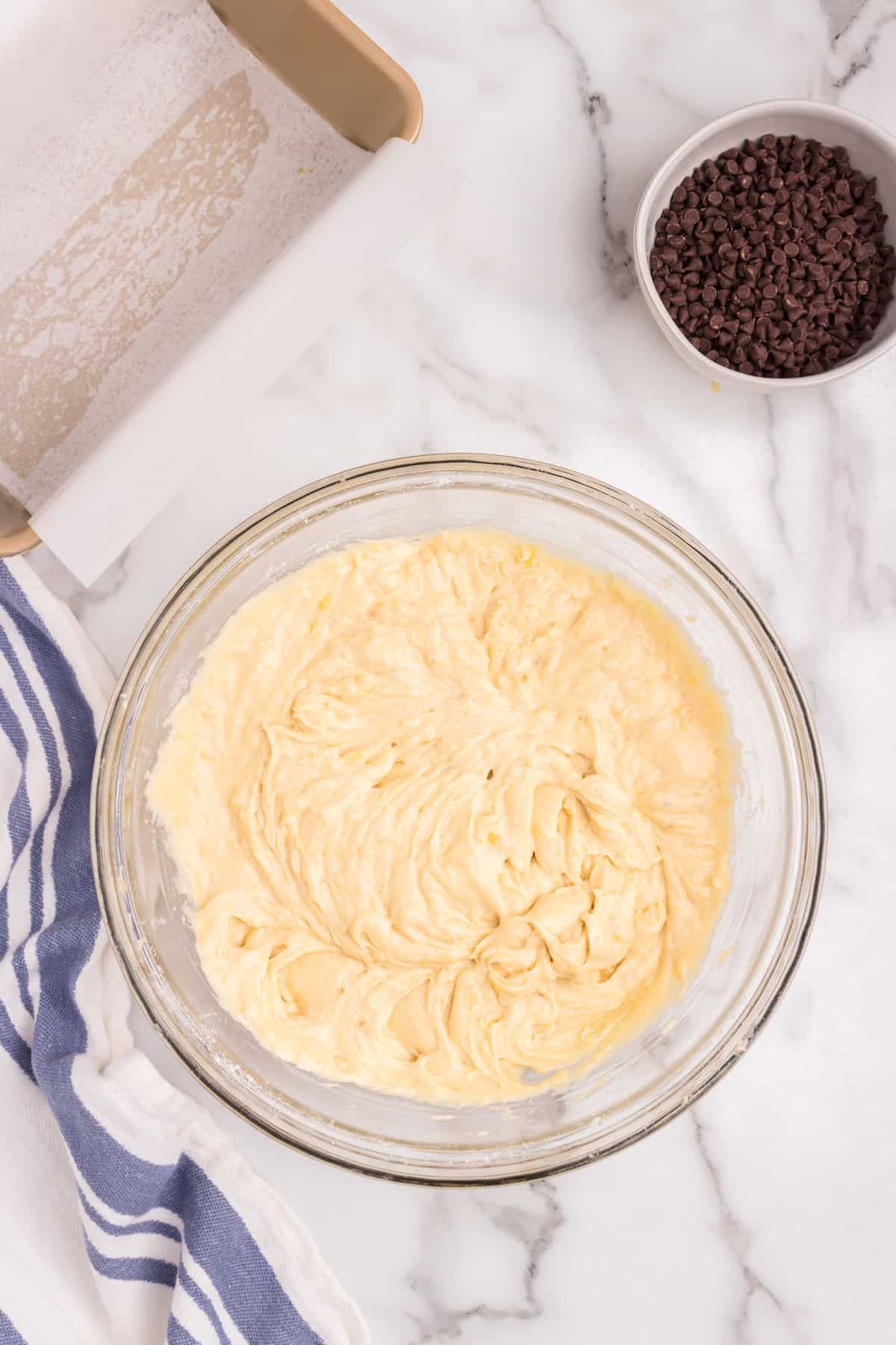 The contents in the mixing bowl are now fully combined into a smooth, creamy banana bread batter. The same parchment-lined loaf pan and bowl of mini chocolate chips remain in the background.