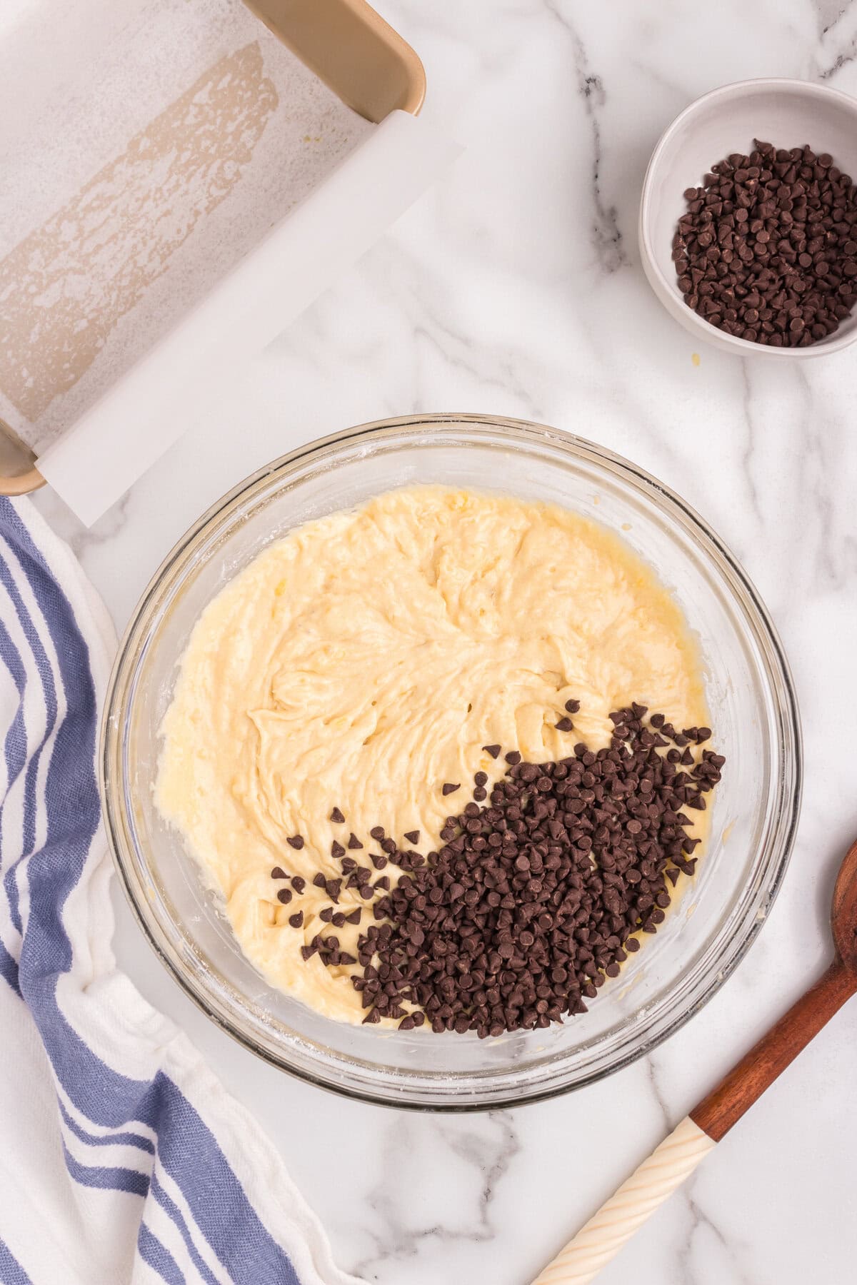 Mini chocolate chips have been added to the bowl of banana bread batter, sitting atop the surface before being mixed in.