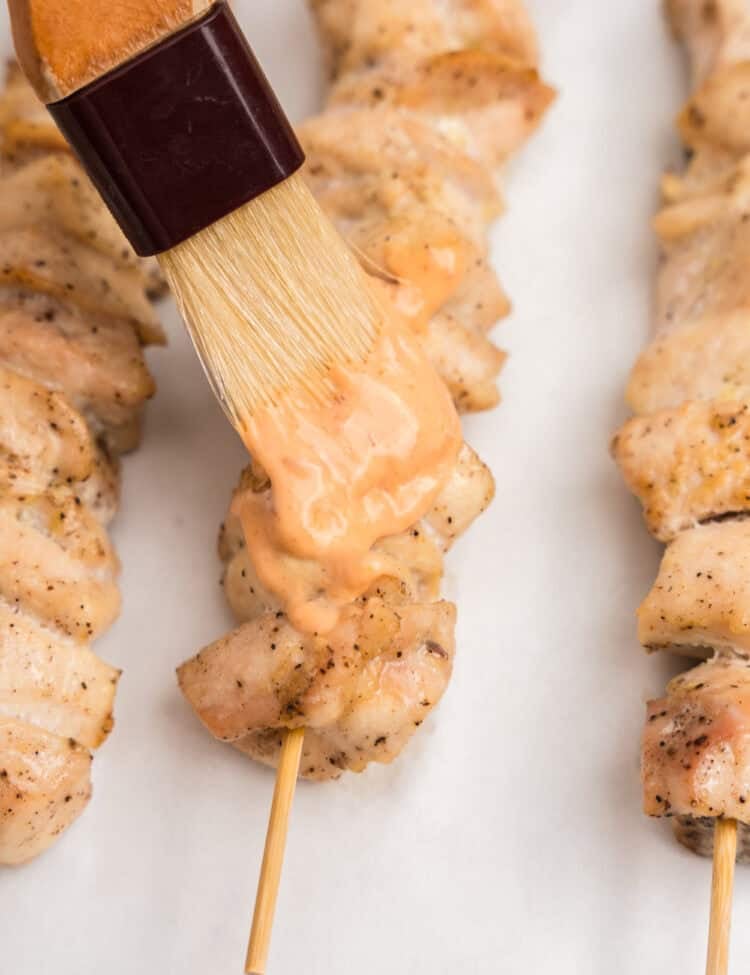 A close-up shot of a pastry brush applying a creamy orange spicy mayo sauce to cooked chicken skewers on parchment paper. The chicken is lightly browned and seasoned.