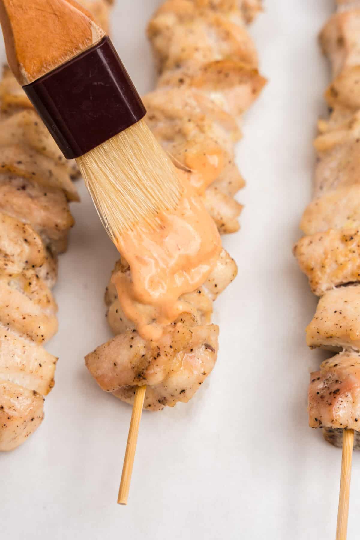 A close-up shot of a pastry brush applying a creamy orange spicy mayo sauce to cooked chicken skewers on parchment paper. The chicken is lightly browned and seasoned.