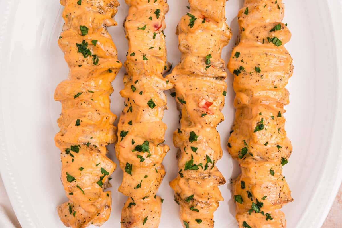 Overhead view of five grilled chicken skewers coated in a spicy orange sauce and garnished with chopped parsley, arranged on a white oval platter.