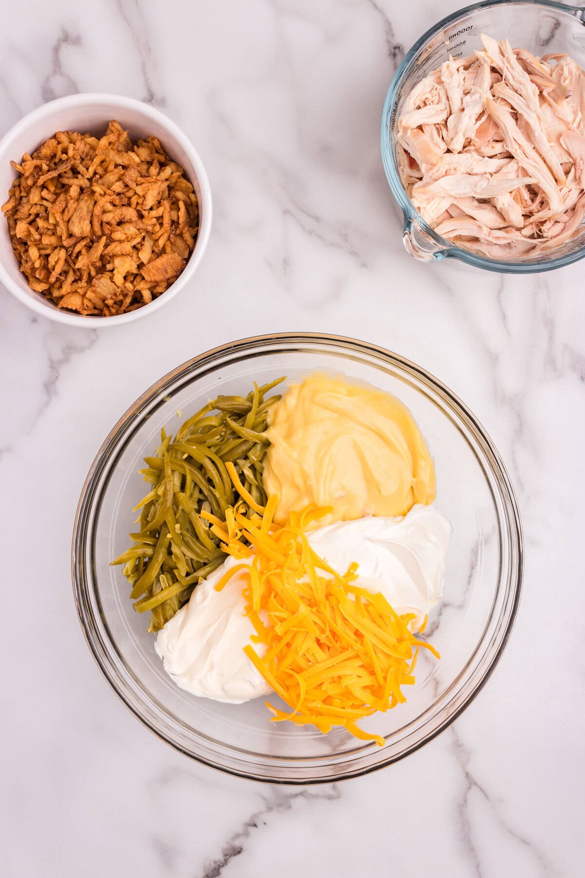 A large glass bowl containing green beans, cream of chicken soup, sour cream, and shredded cheddar cheese, with separate bowls of shredded chicken and crispy fried onions nearby.