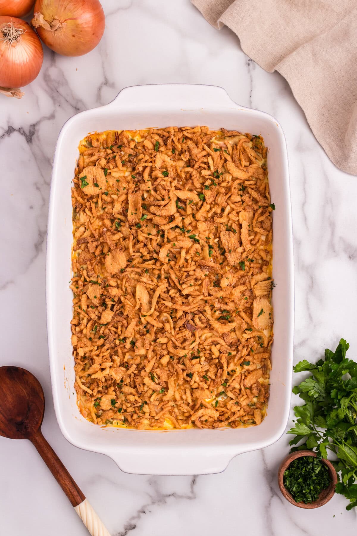 Freshly baked French onion chicken casserole topped with crispy fried onions and sprinkled with chopped parsley in a white baking dish, surrounded by onions, herbs, and a wooden spoon.