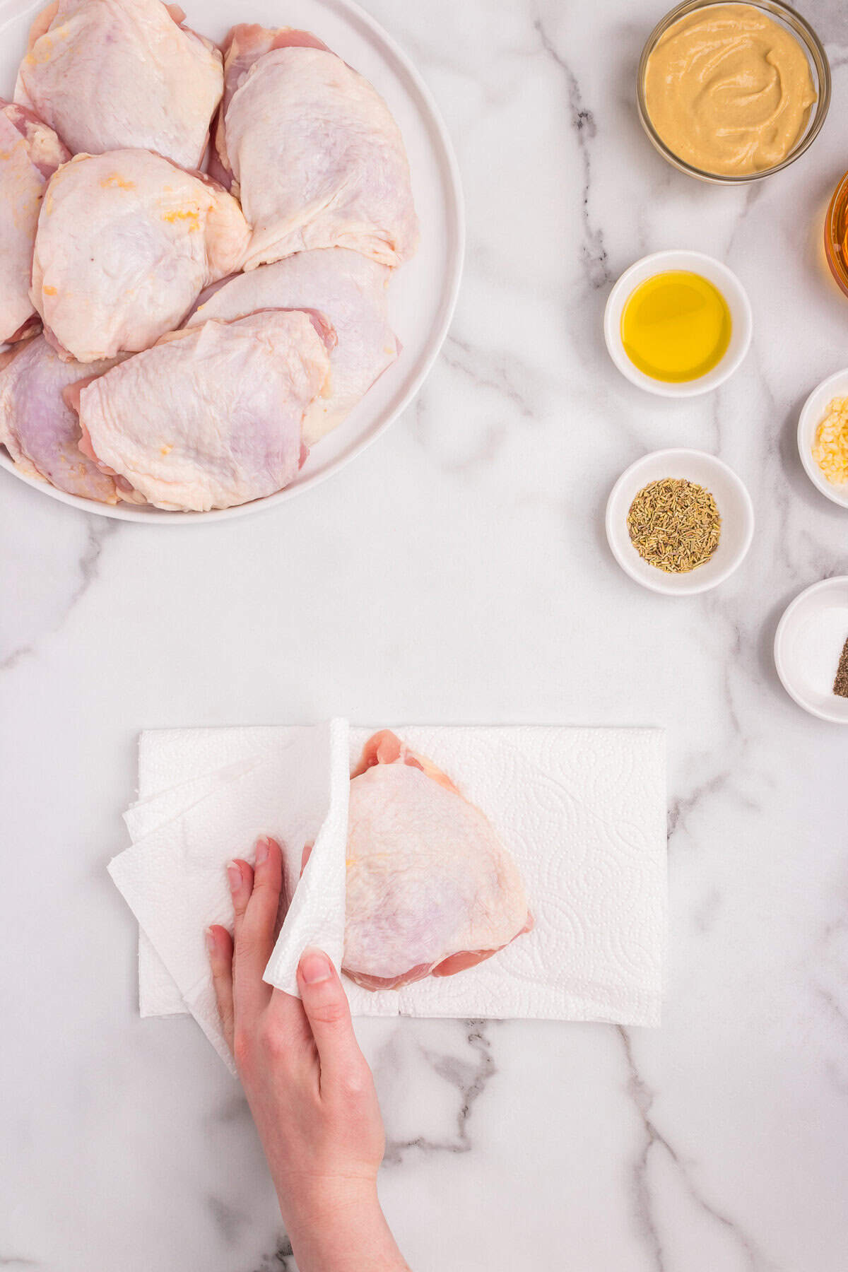 A person patting raw chicken thighs dry with paper towels on a marble countertop.