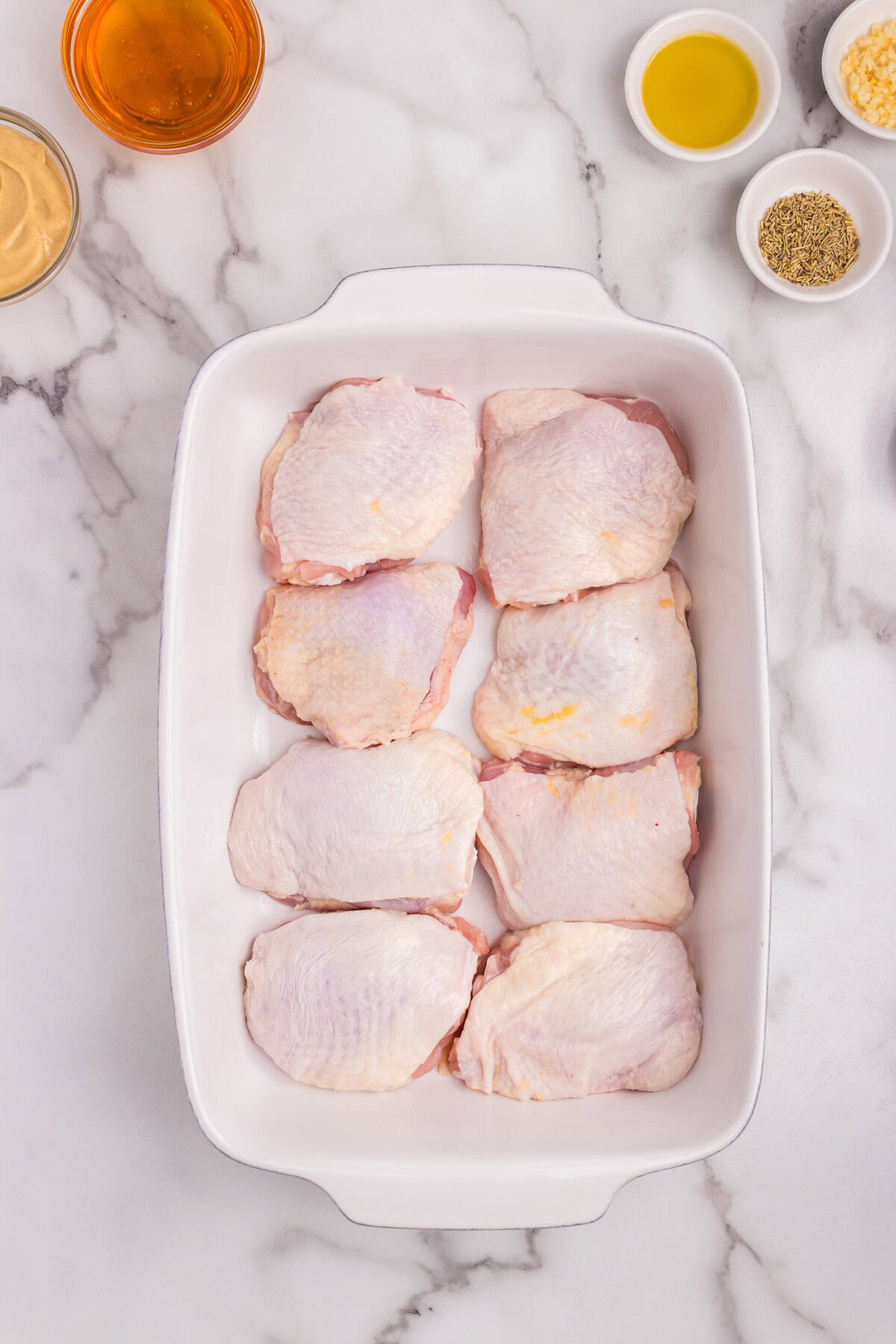 Eight raw chicken thighs arranged snugly in a white ceramic baking dish.