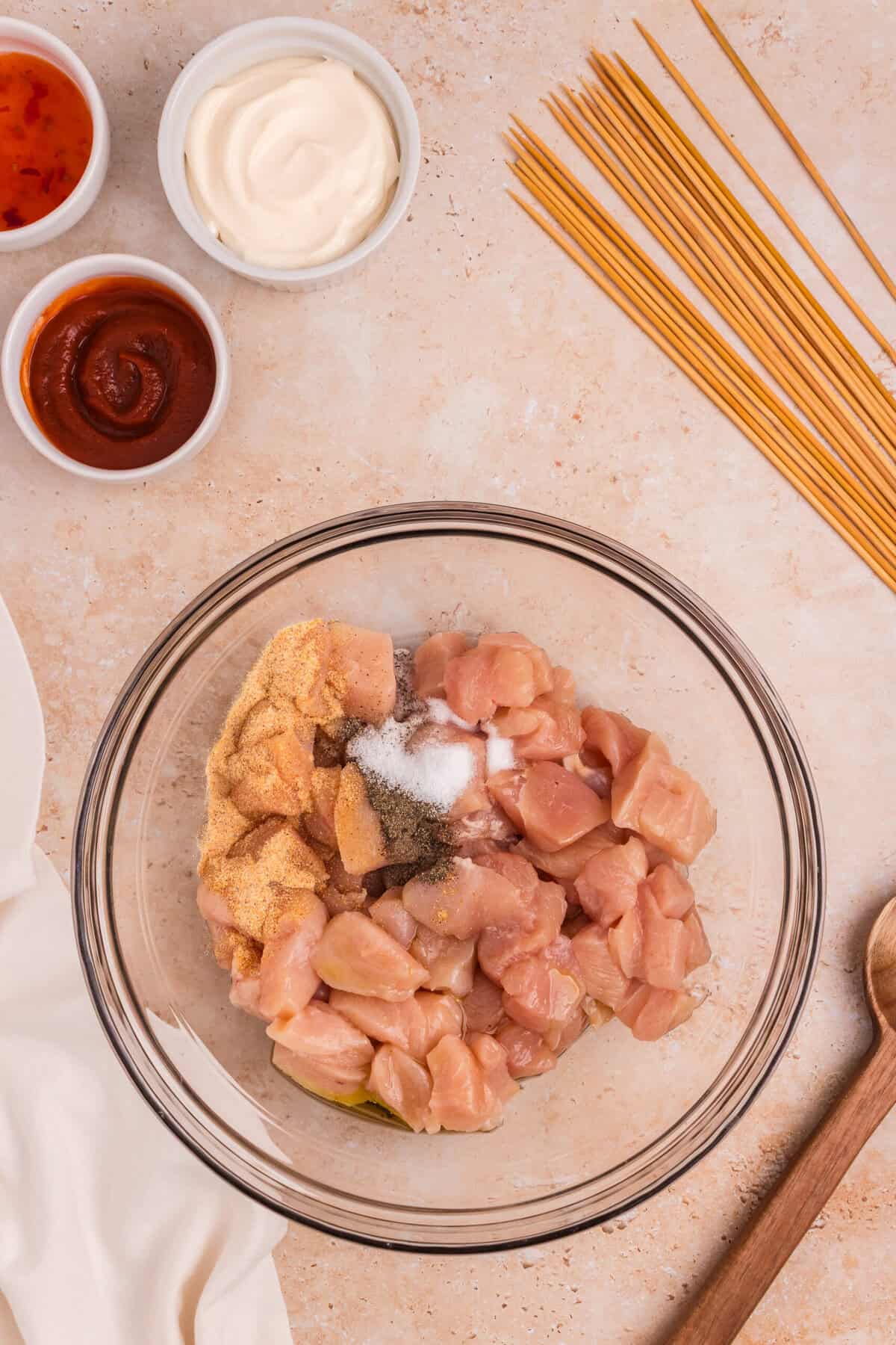 A bowl of raw diced chicken breast with seasoning on top (including garlic powder, salt, black pepper, and oil) on a beige surface, surrounded by wooden skewers and small bowls of mayonnaise, sweet chili sauce, and sriracha sauce.