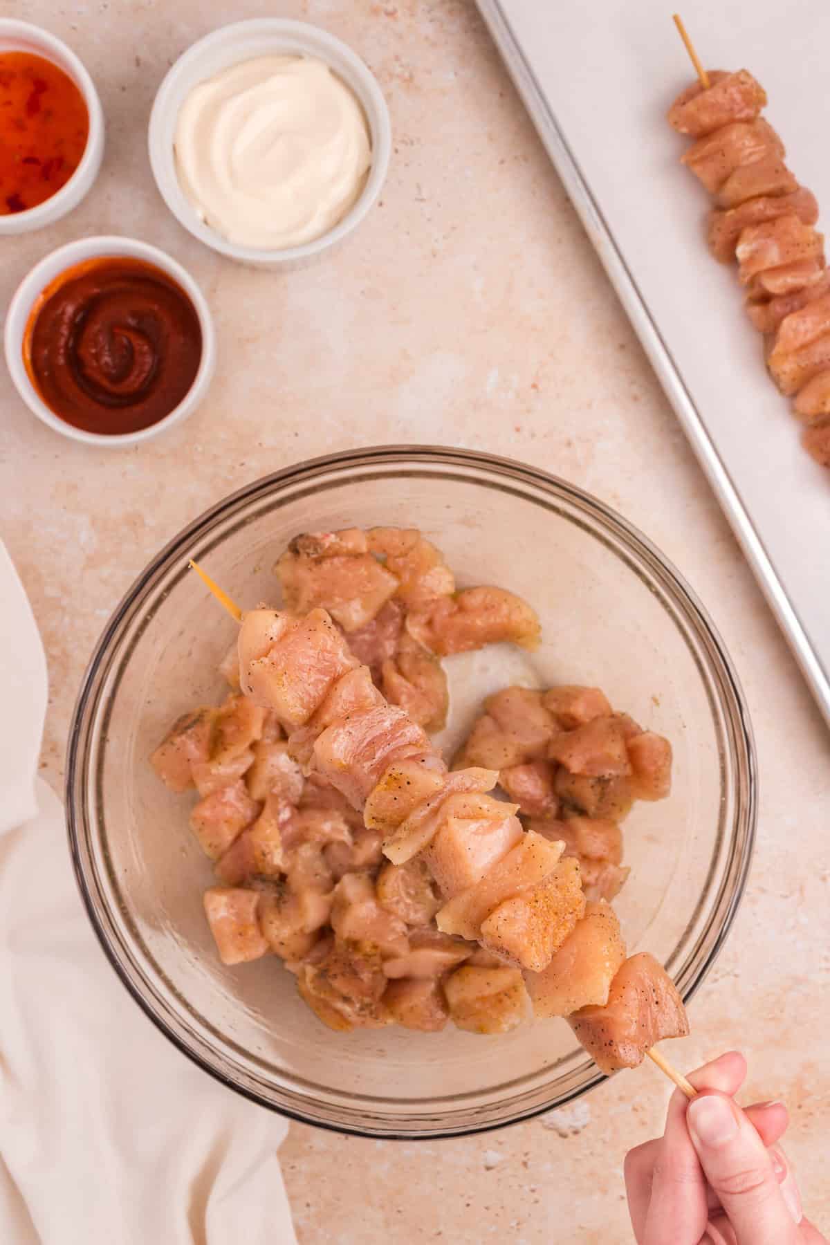 A hand holding a wooden skewer being threaded with the seasoned chicken pieces over the bowl. A tray with already skewered chicken is partially visible in the background.