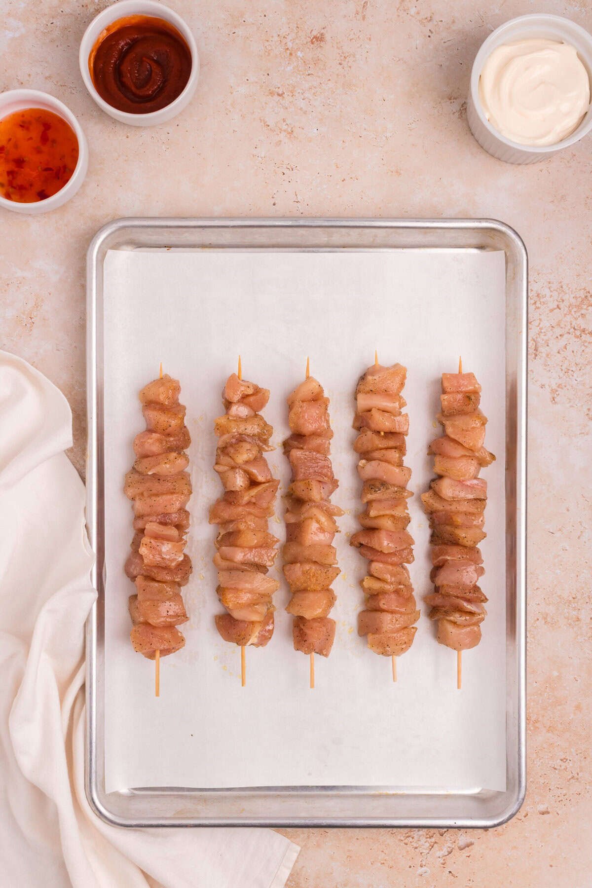A baking tray lined with parchment paper holding six uncooked chicken skewers, neatly arranged in rows, ready to be cooked. Bowls of sauce and skewers remain visible in the background.