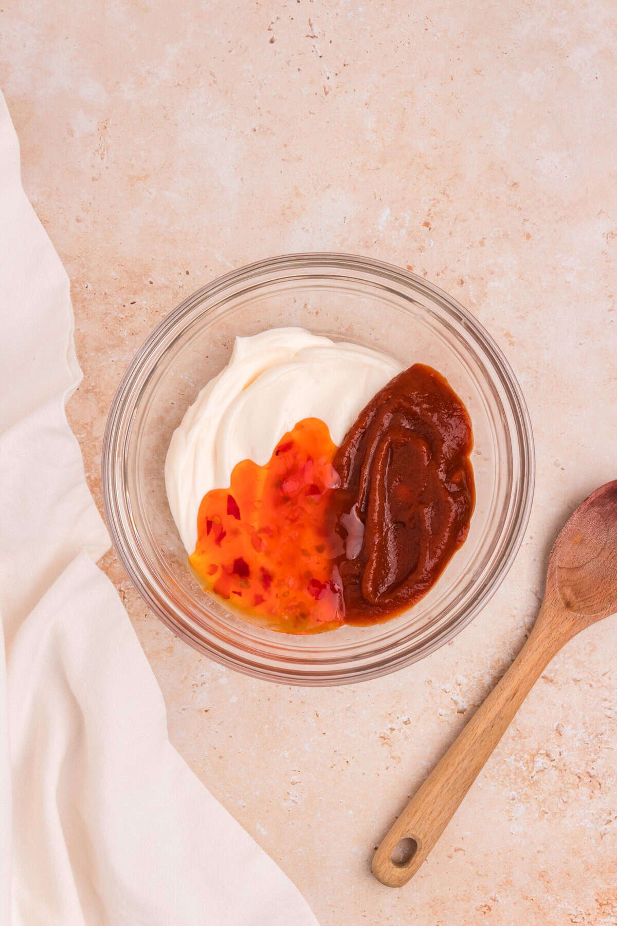 A glass bowl containing mayonnaise, sweet chili sauce, and sriracha sauce side by side, before being mixed into a dipping sauce. A wooden spoon lies nearby.