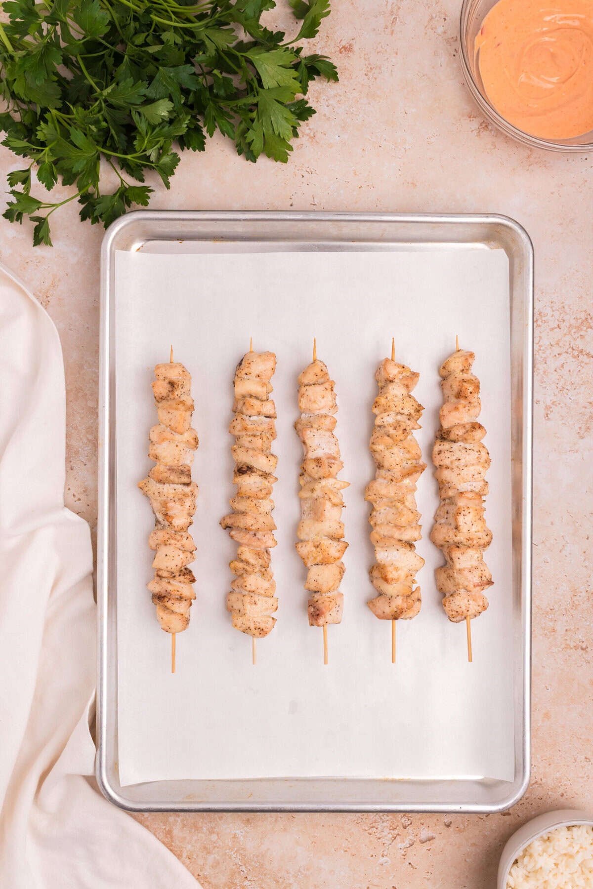 The chicken skewers have been baked and are now fully cooked, lightly browned and resting on the parchment-lined tray. Fresh parsley and dipping sauce are seen nearby.