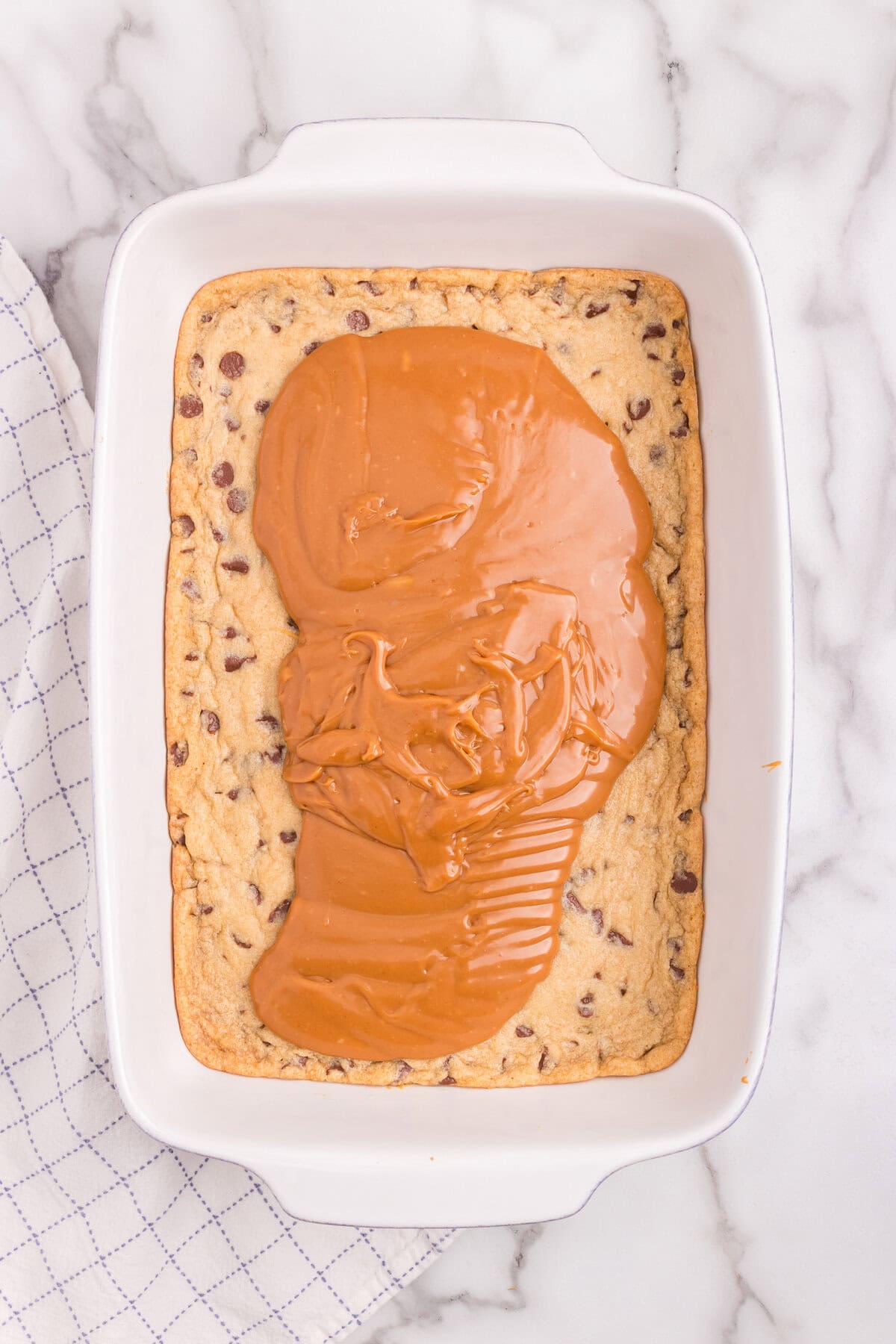 Melted caramel is being spread over the baked chocolate chip cookie layer in the baking dish.
