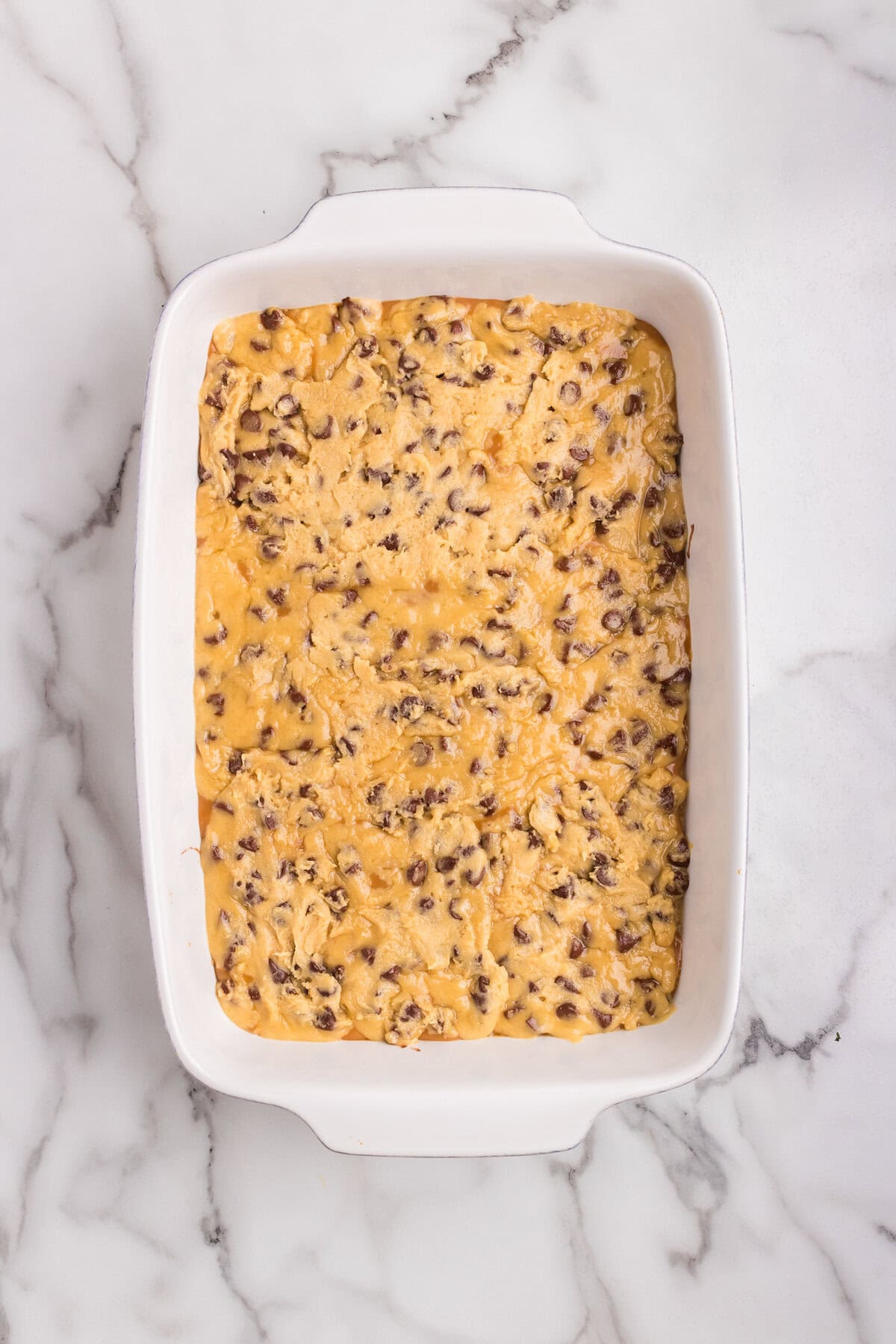 The second layer of chocolate chip cookie dough is spread over the caramel layer in a white baking dish, ready to be baked.