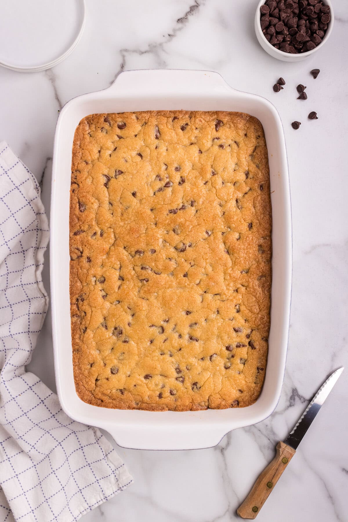 A wider shot of the baked bars in the dish, resting on a marble counter with a knife and a bowl of chocolate chips nearby.