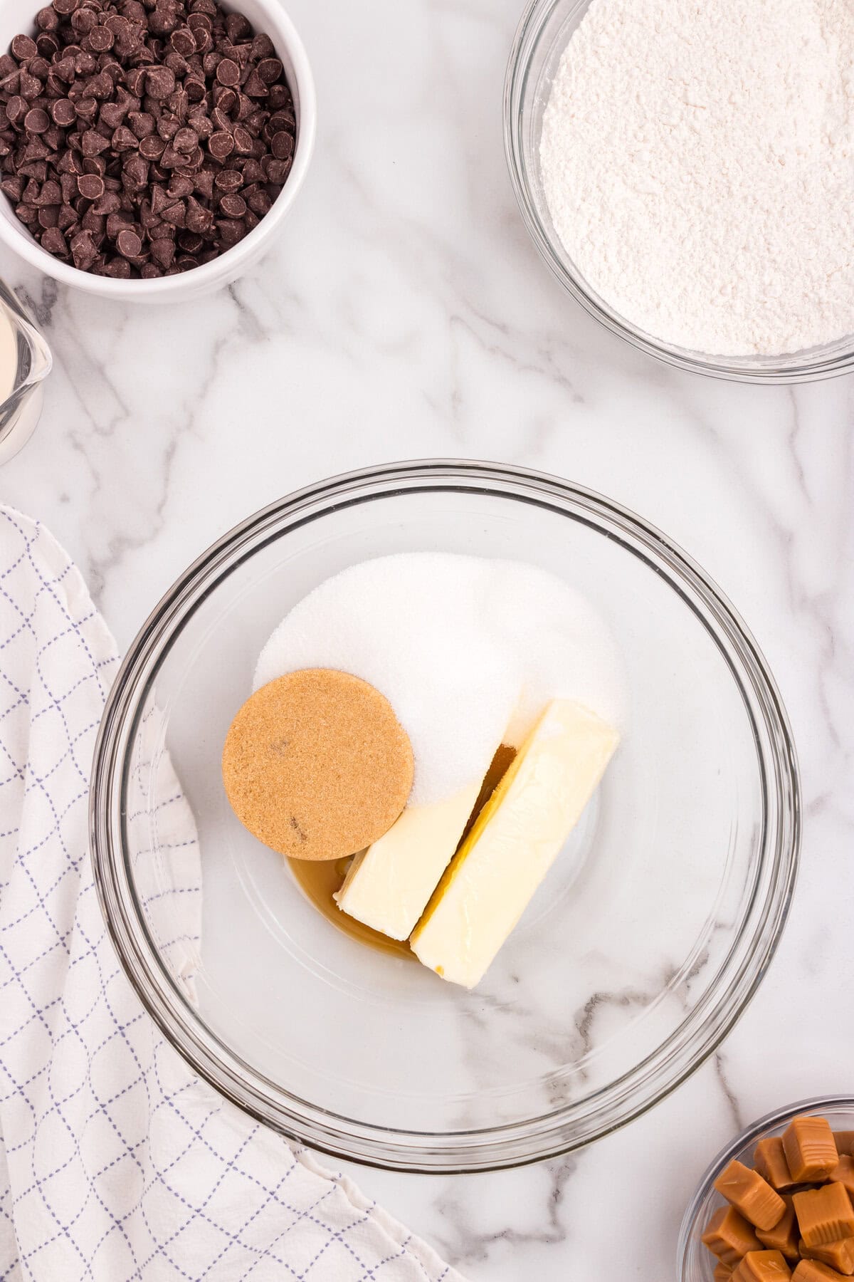 A glass mixing bowl with softened butter, brown sugar, and granulated sugar ready to be creamed together. Surrounding bowls contain chocolate chips, flour, and caramels.