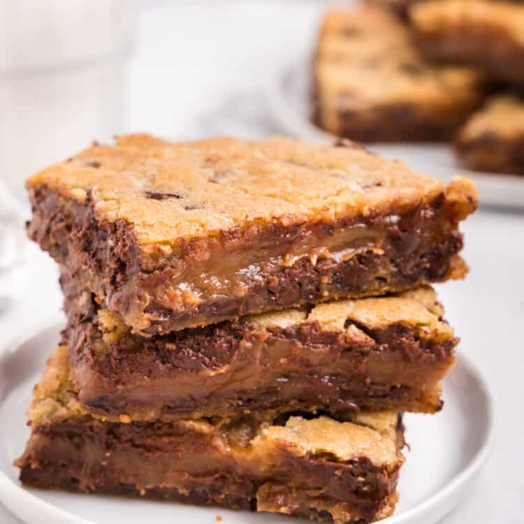 A stack of three gooey caramel chocolate chip bars on a white plate, with a glass of milk and more bars in the background.