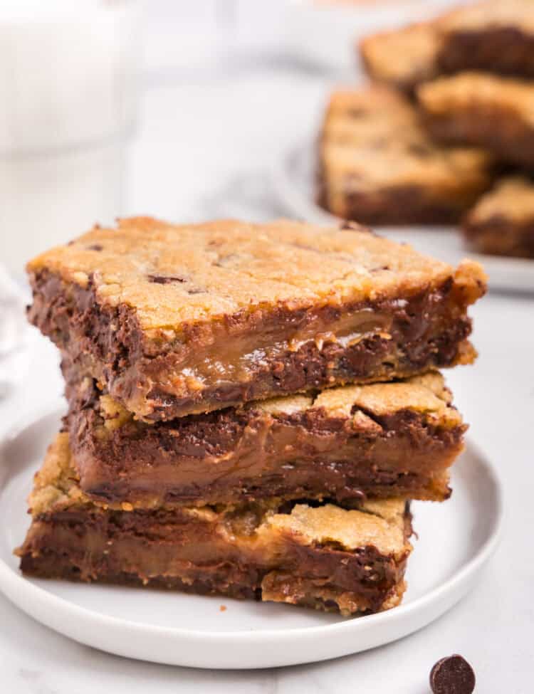 A stack of three gooey caramel chocolate chip bars on a white plate, with a glass of milk and more bars in the background.