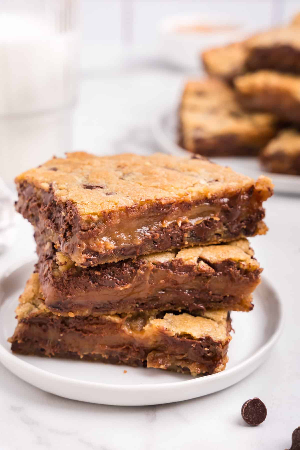 A stack of three gooey caramel chocolate chip bars on a white plate, with a glass of milk and more bars in the background.