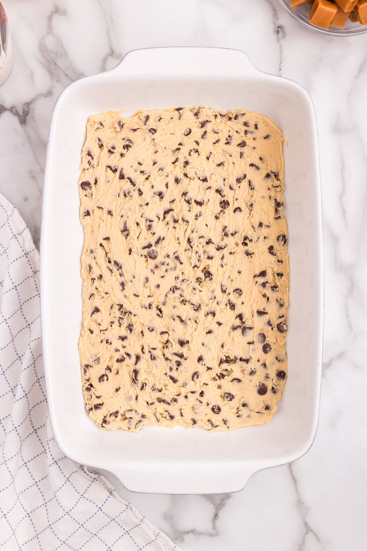 The chocolate chip cookie dough is spread evenly into a rectangular white baking dish, forming the base layer.