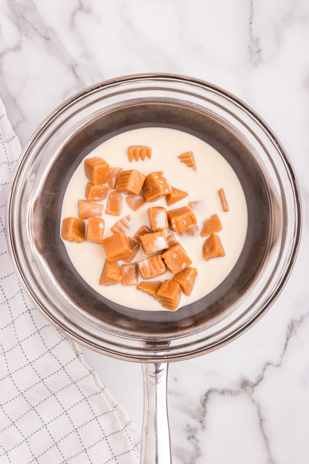 A heatproof glass bowl over a saucepan (double boiler setup) with caramel candies and evaporated milk, beginning to melt.