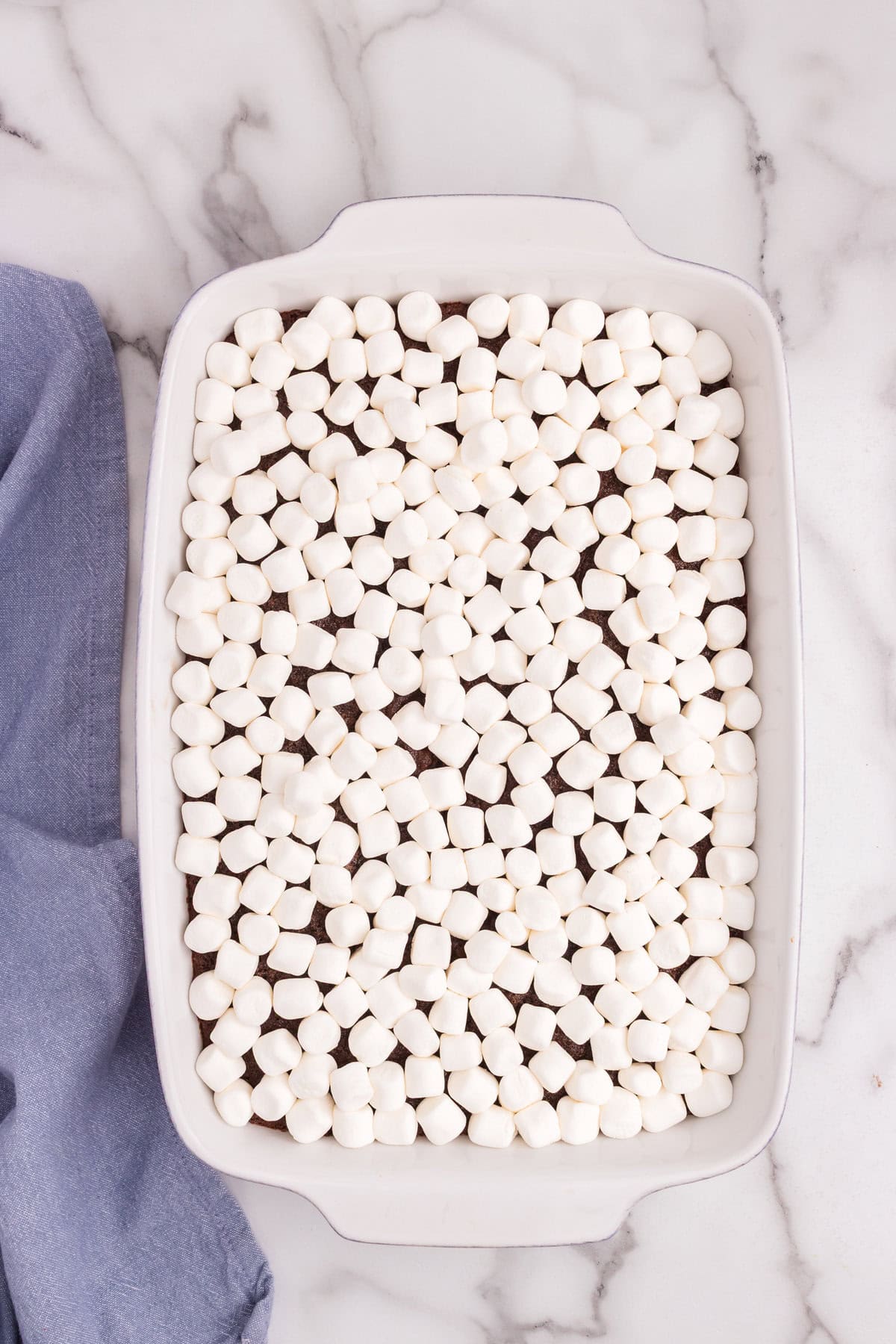 Baked chocolate cake topped with a full layer of mini marshmallows in a white rectangular baking dish, ready to be melted.