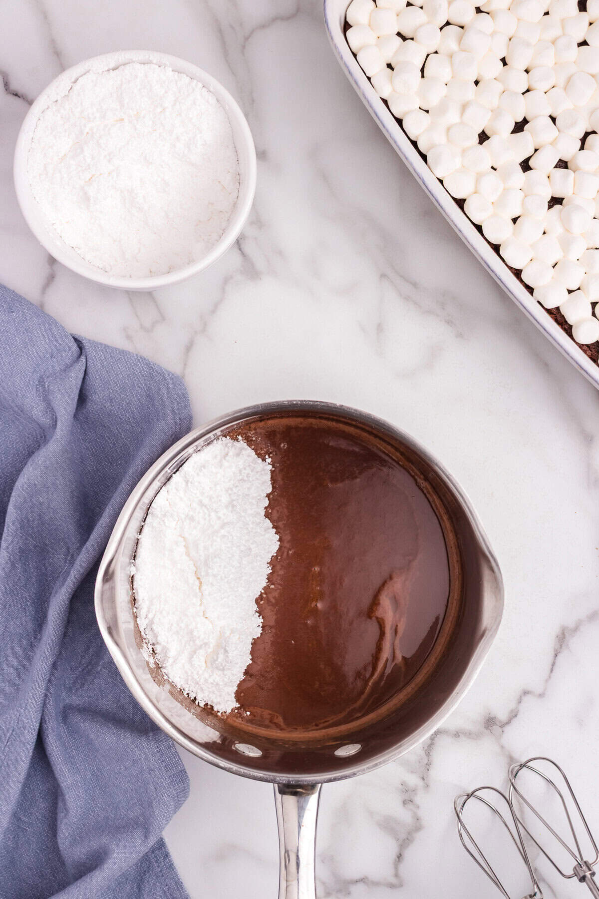 Powdered sugar and chocolate sauce being combined in a saucepan, with the marshmallow-topped cake visible nearby.