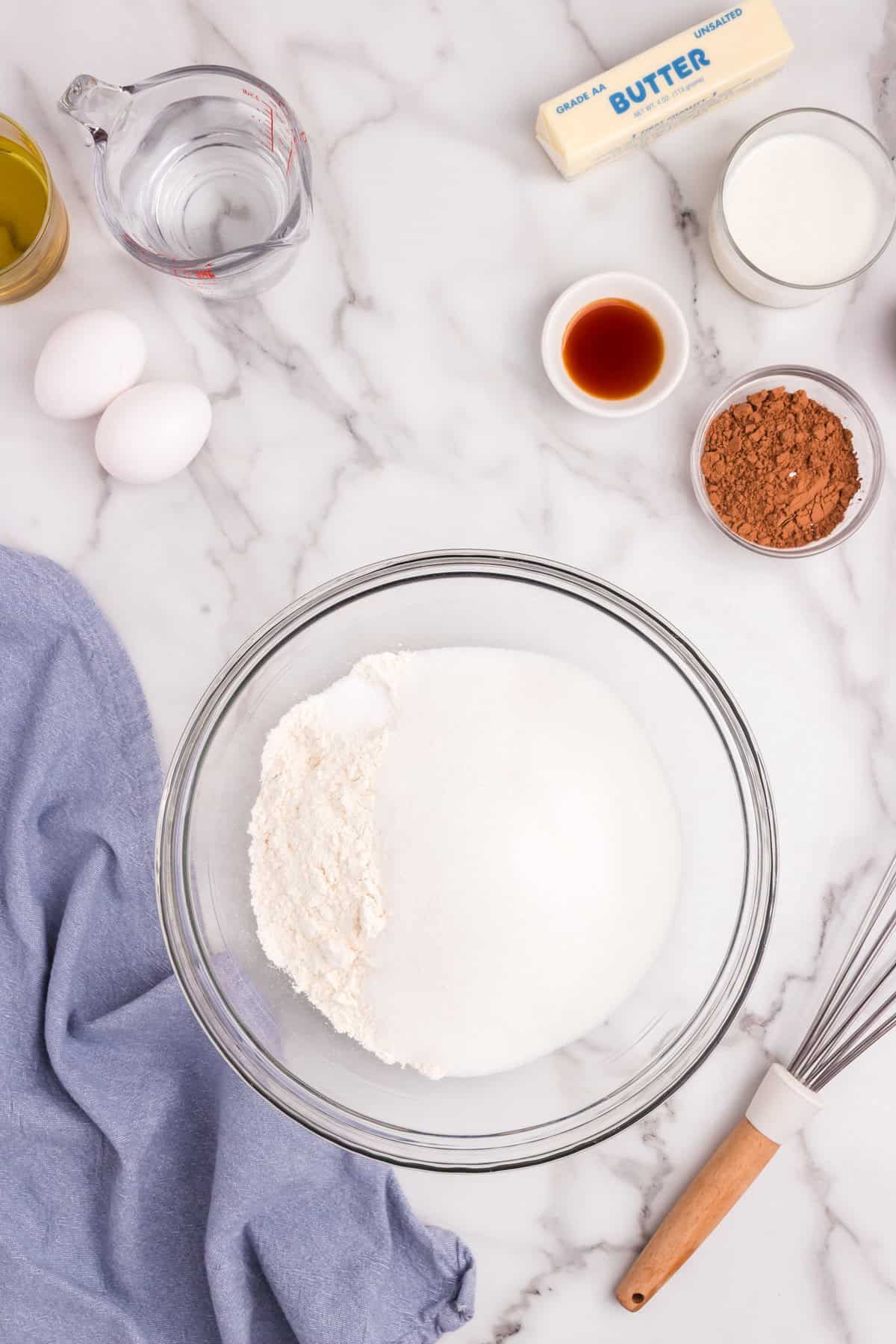 Overhead view of baking ingredients on a marble counter: a bowl of flour and sugar, eggs, milk, vanilla extract, cocoa powder, butter, vegetable oil, and water.