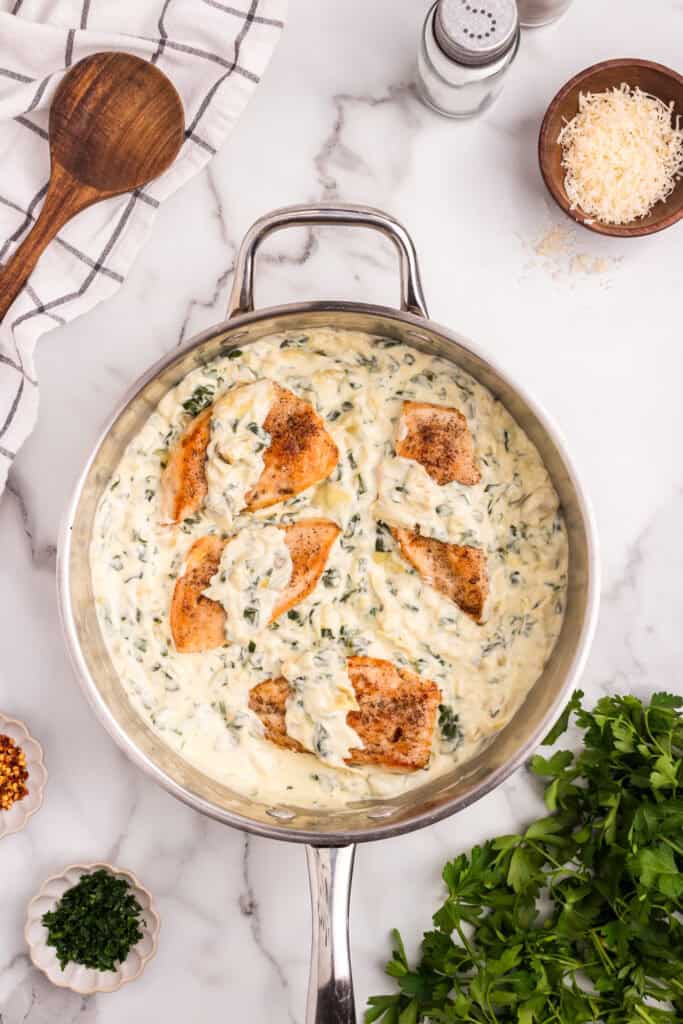 Top-down view of a stainless steel skillet filled with seared chicken breasts smothered in a creamy spinach and artichoke sauce, surrounded by garnishes including shredded cheese, parsley, red pepper flakes, and a wooden spoon on a marble countertop.