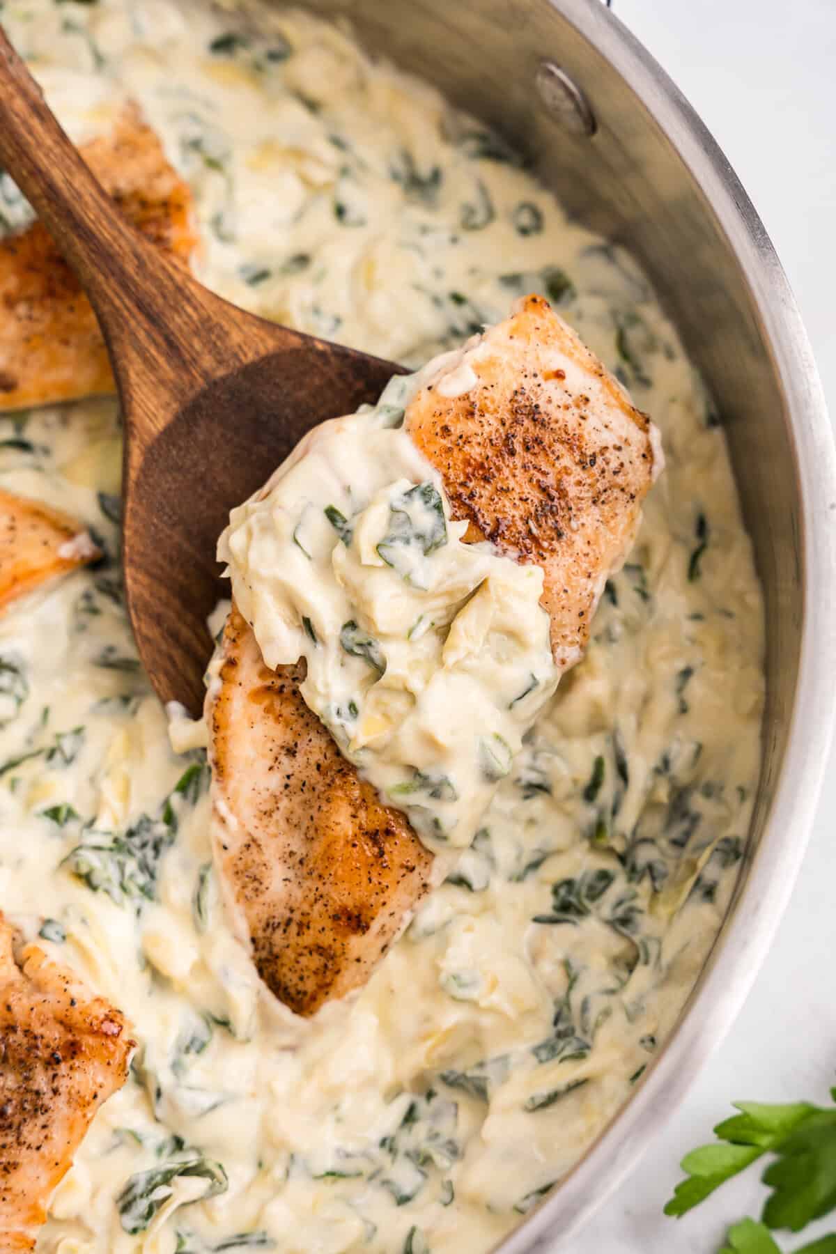 A close-up of a creamy spinach artichoke chicken dish in a stainless steel skillet. A wooden spoon holds a piece of seared, seasoned chicken breast topped with a rich, cheesy spinach and artichoke sauce. The sauce is thick and creamy, generously coating the chicken and surrounding the skillet.