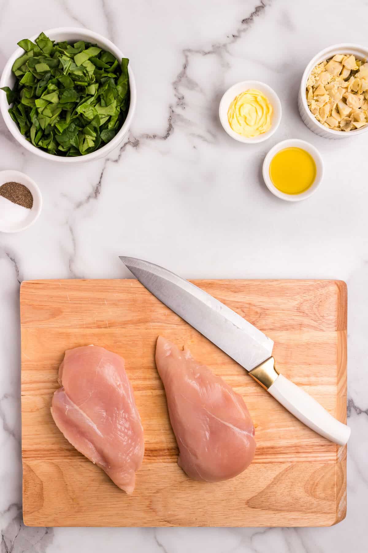 Ingredients laid out on a marble surface including raw chicken breasts on a wooden cutting board, chopped spinach, artichoke hearts, olive oil, butter, salt, and pepper.