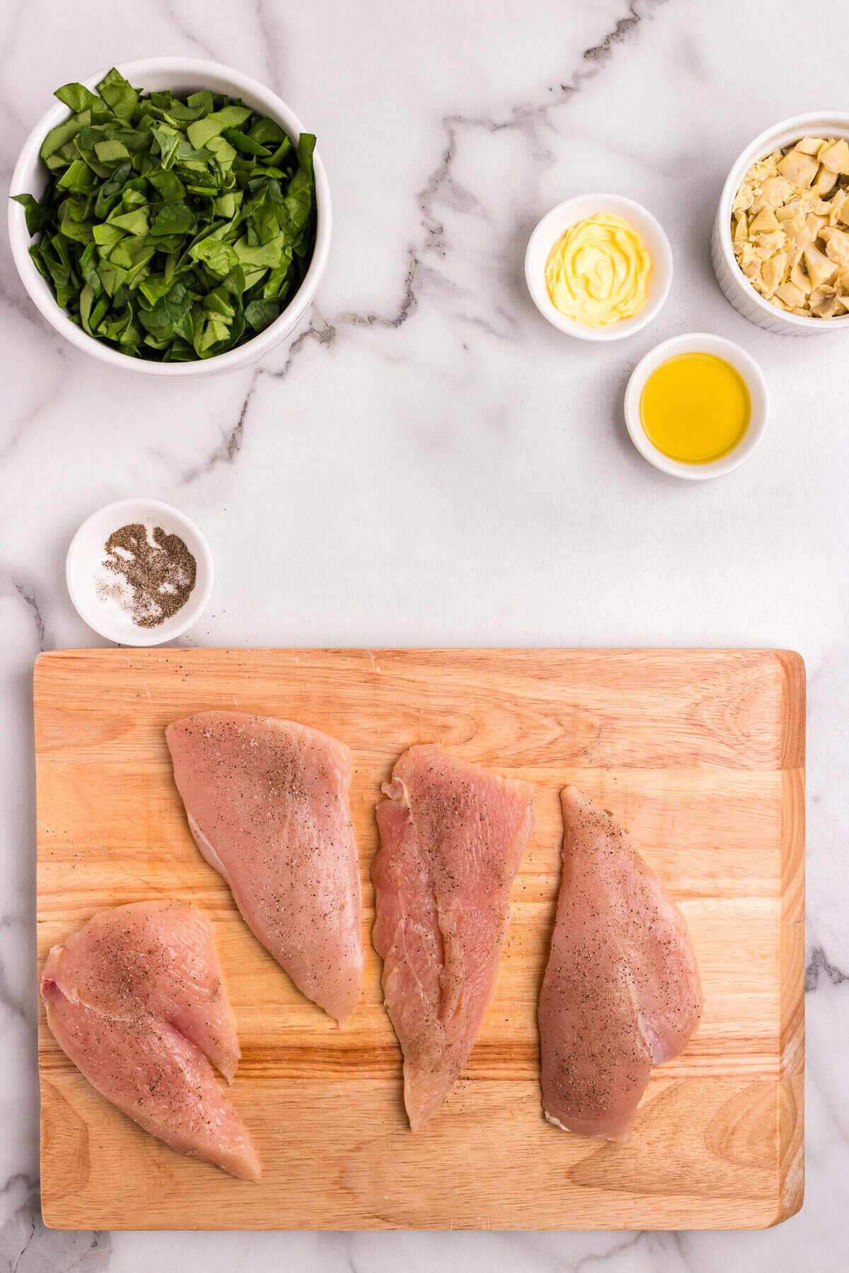 Chicken breasts sliced and seasoned with salt and pepper, displayed on a wooden cutting board next to bowls of chopped spinach, artichoke hearts, olive oil, and butter.