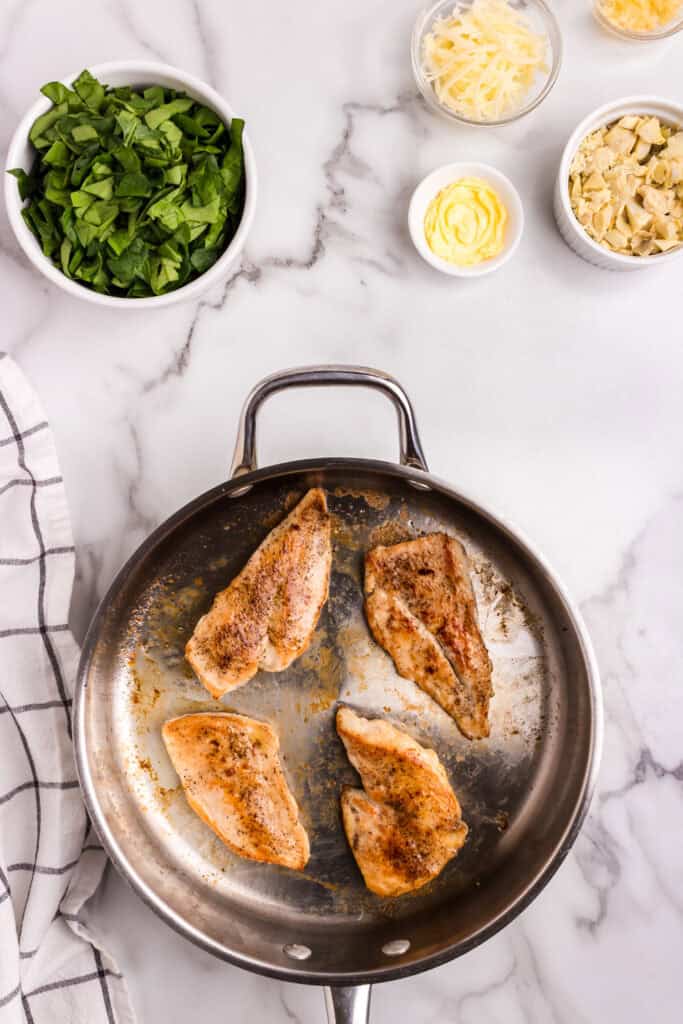 Seasoned chicken breasts cooking in a stainless steel skillet, with bowls of chopped spinach, shredded cheese, artichoke hearts, and butter nearby.