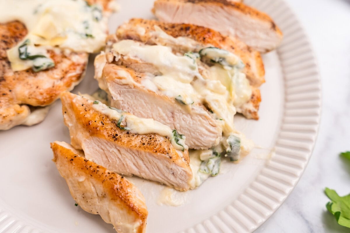 Seared chicken breasts simmered in a creamy, cheesy spinach and artichoke sauce sliced on a white plate.