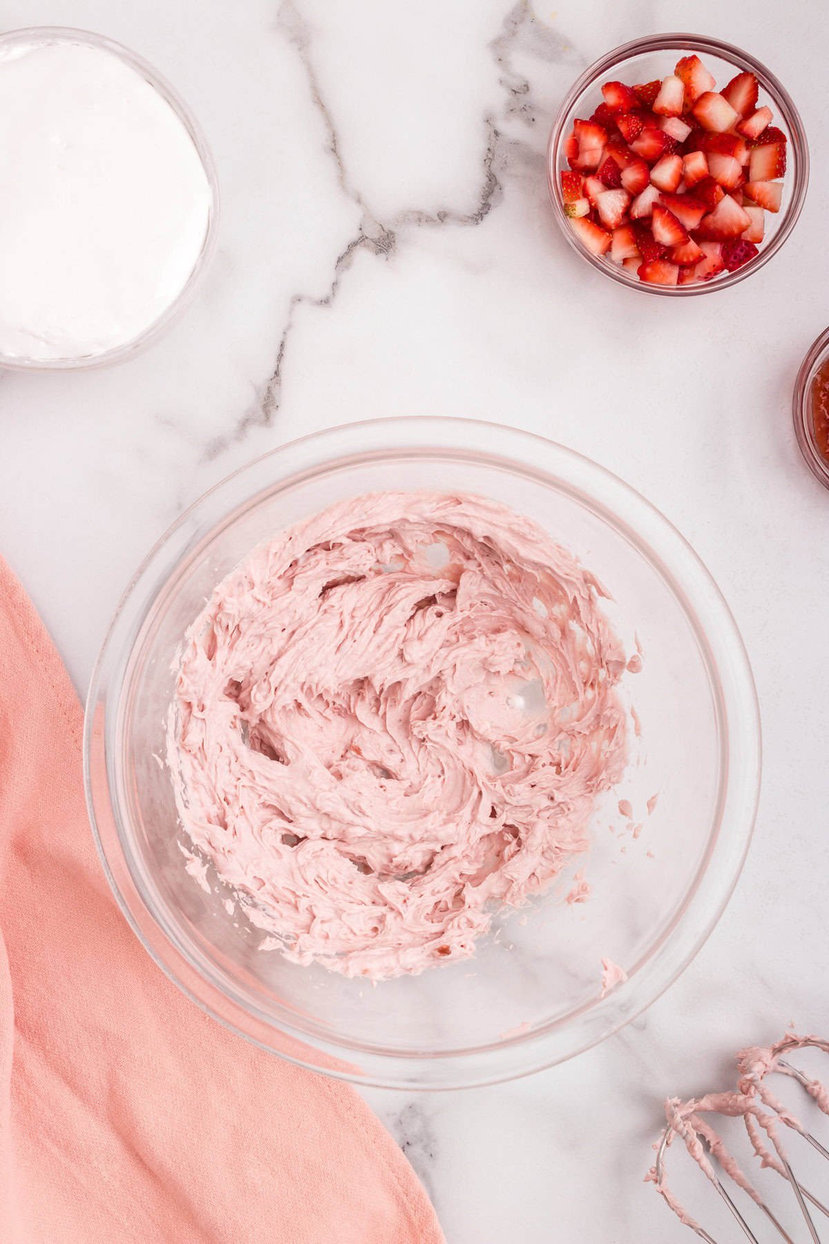 A glass bowl filled with pink whipped strawberry mixture sitting on a white marble surface. Surrounding the bowl are small bowls of whipped topping, chopped strawberries, and strawberry preserves.