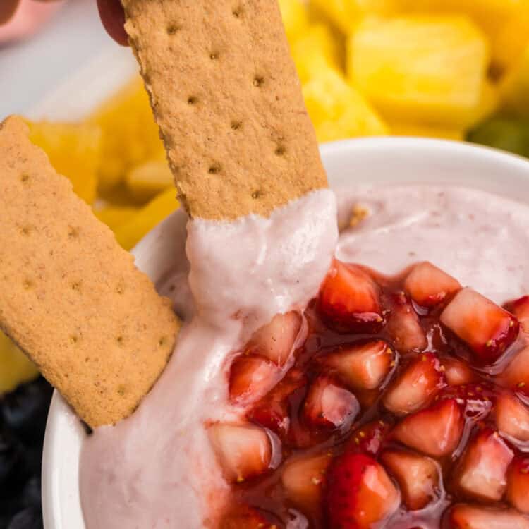 A close-up of a hand dipping a rectangular graham cracker into the creamy pink strawberry dip, topped with shiny chopped strawberries.
