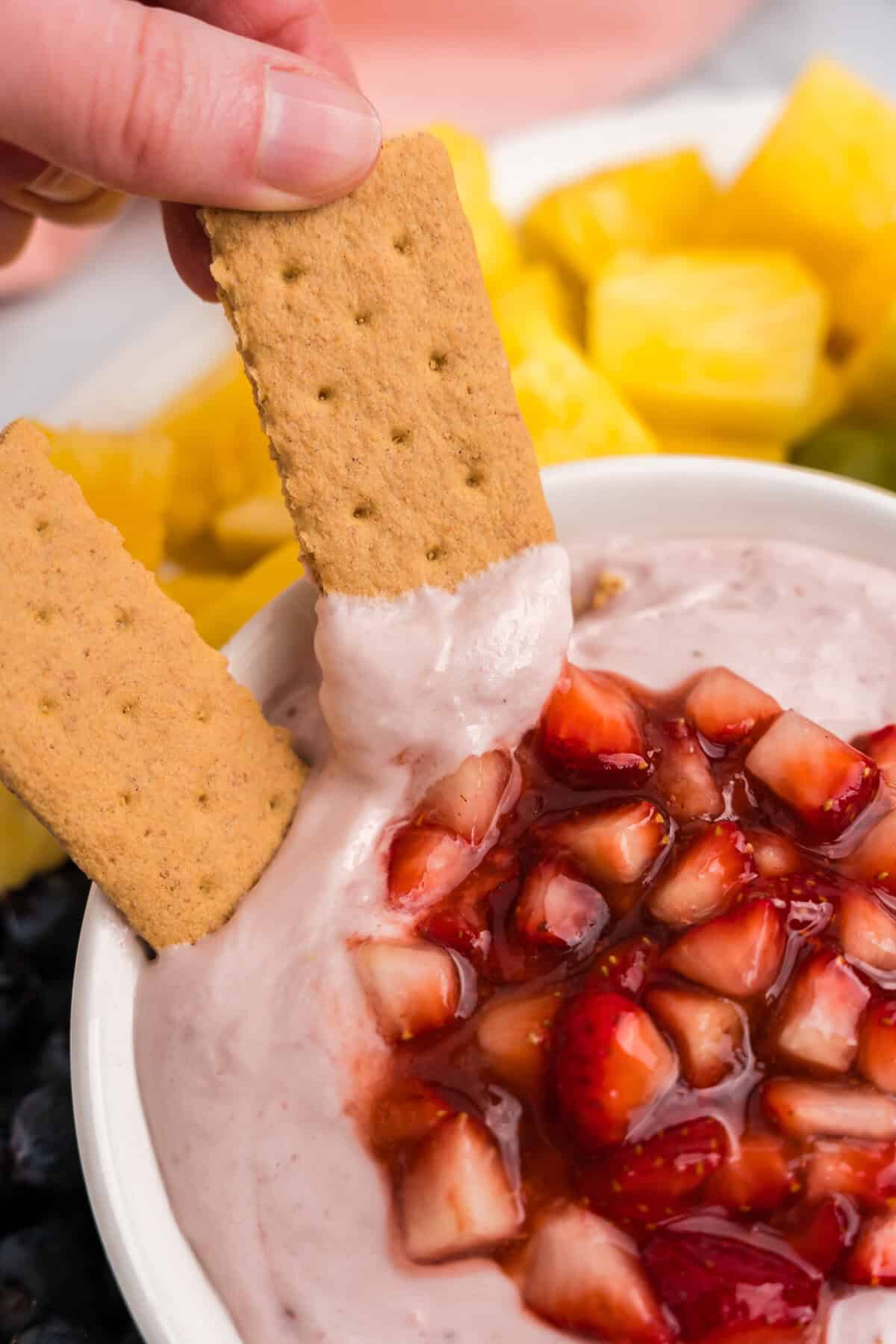 A close-up of a hand dipping a rectangular graham cracker into the creamy pink strawberry dip, topped with shiny chopped strawberries.