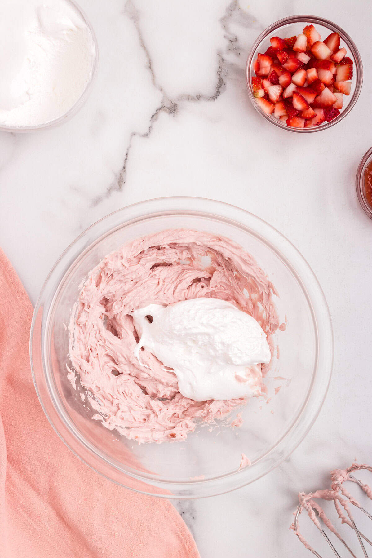 A large dollop of white whipped topping is added to the pink strawberry mixture in a glass bowl, ready to be folded together. Other ingredients like chopped strawberries and preserves are nearby.