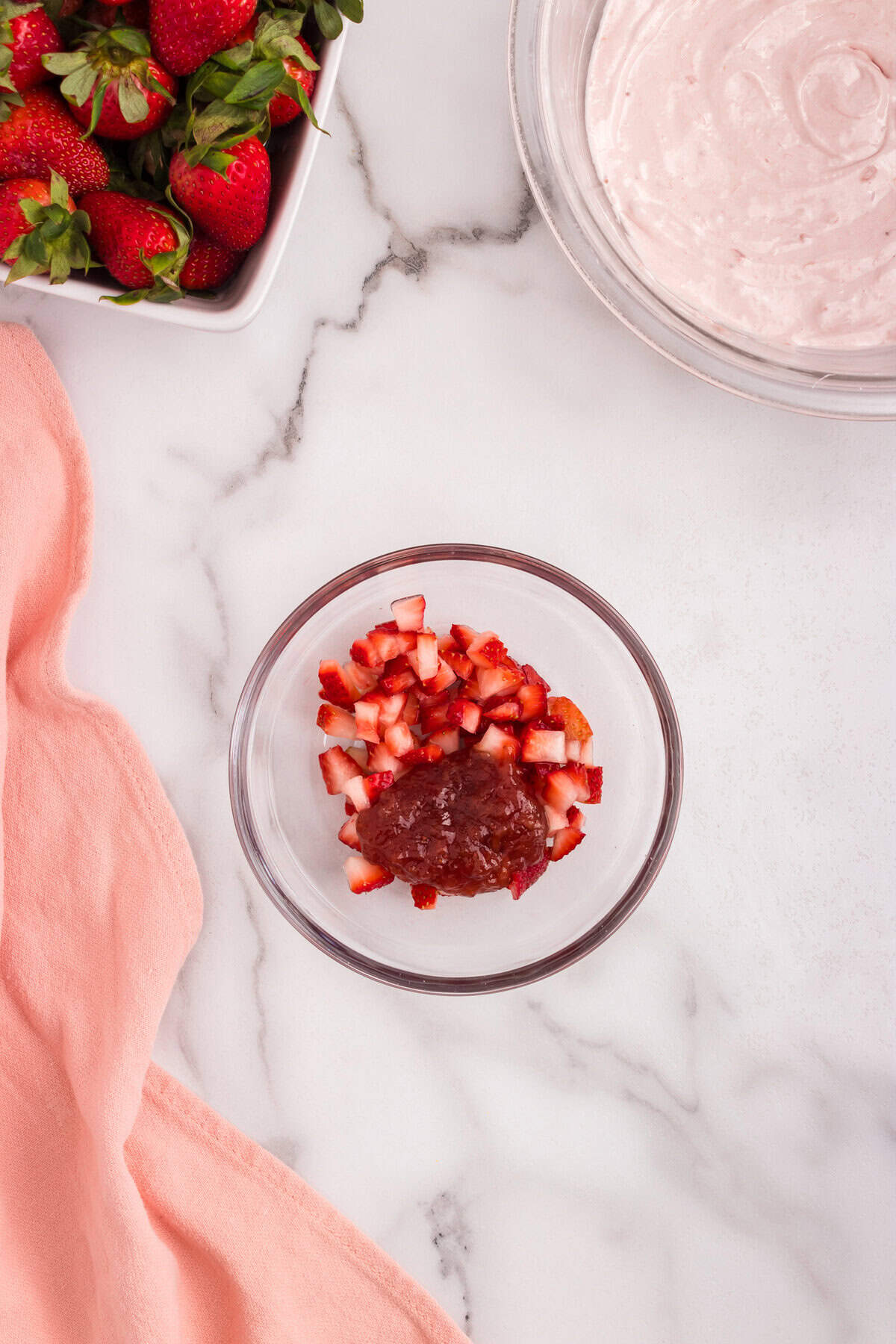 A small glass bowl containing chopped strawberries and a scoop of strawberry preserves, sitting on a marble counter next to a pink cloth.